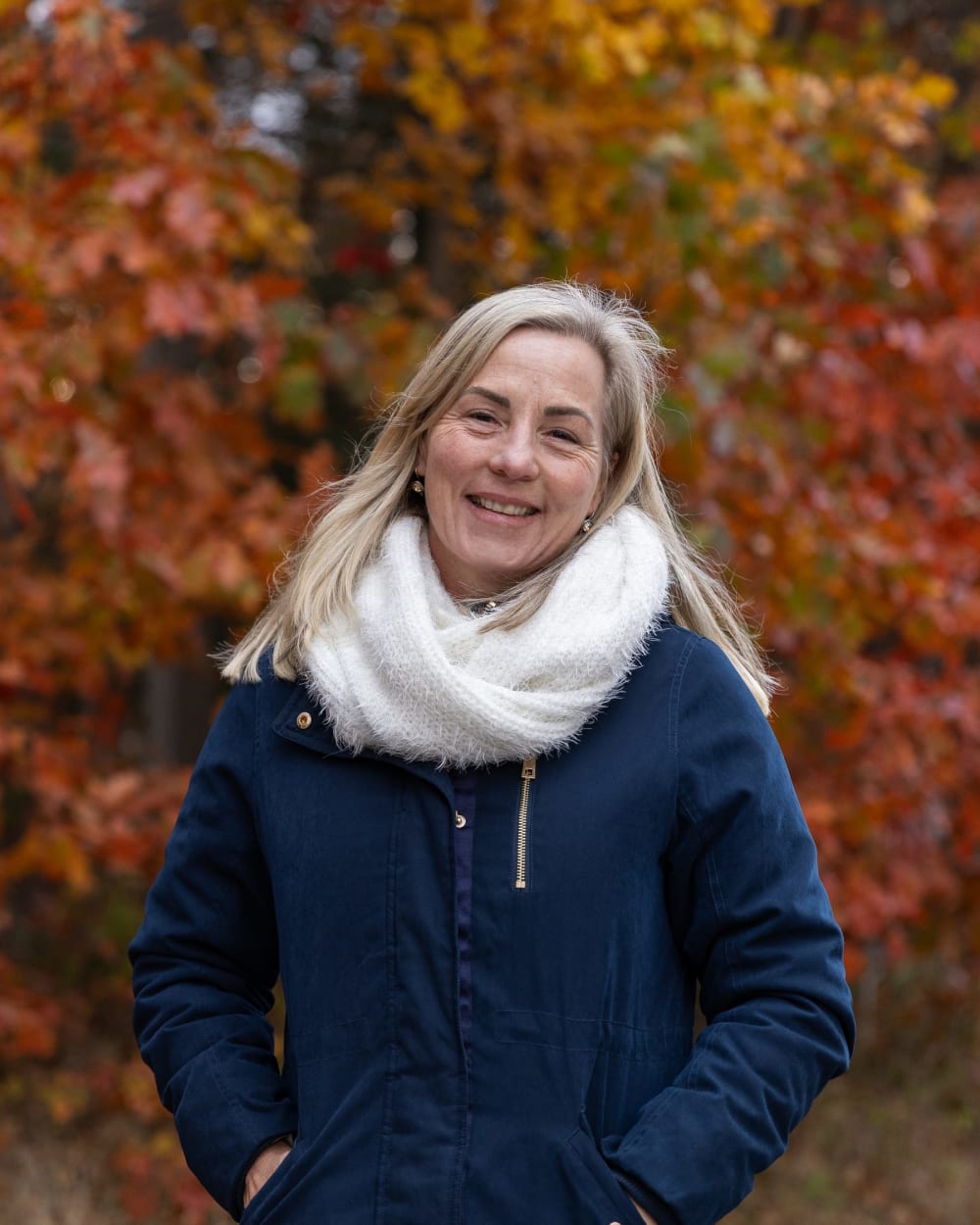A woman in a navy coat and cream scarf smiling in front of red leaves