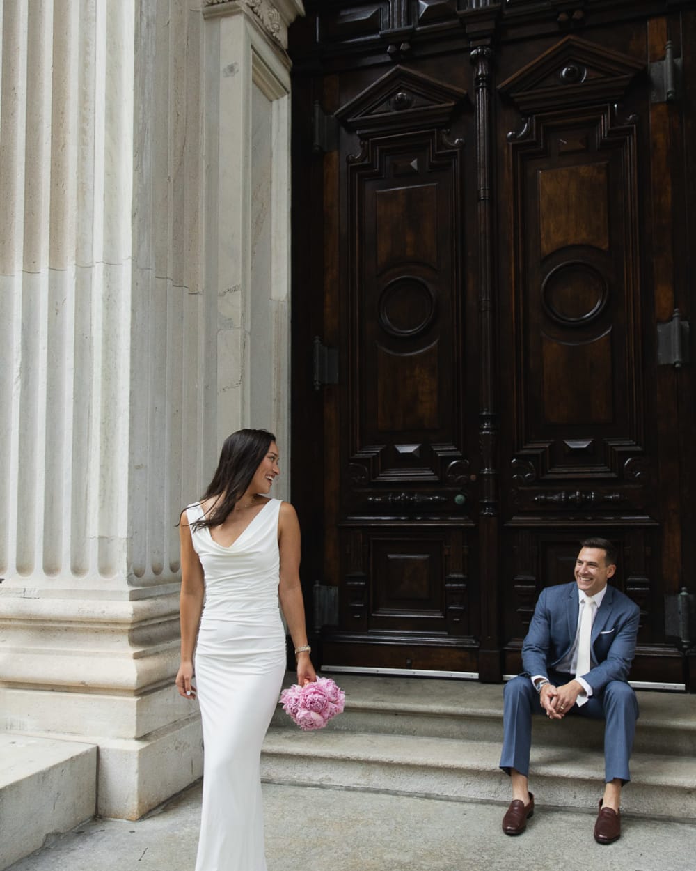 Bride walking past the columns with her peony bouquet while the groom sits laughing on the steps
