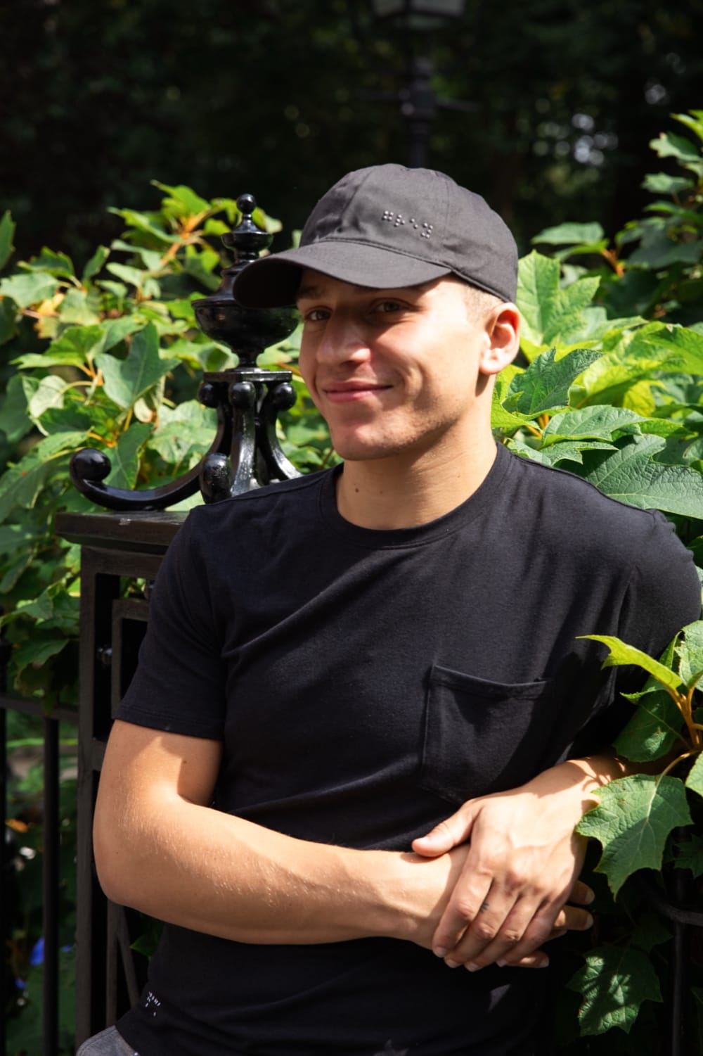 A model in a black cap and tee leaning against a park fence in a lifestyle campaign in New York