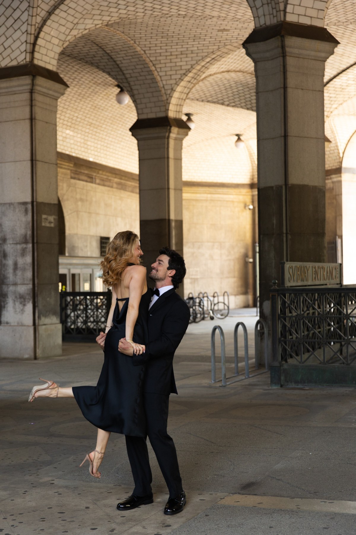 A groom lifting his bride as they laugh under the vaulted stone arcade by City Hall in New York
