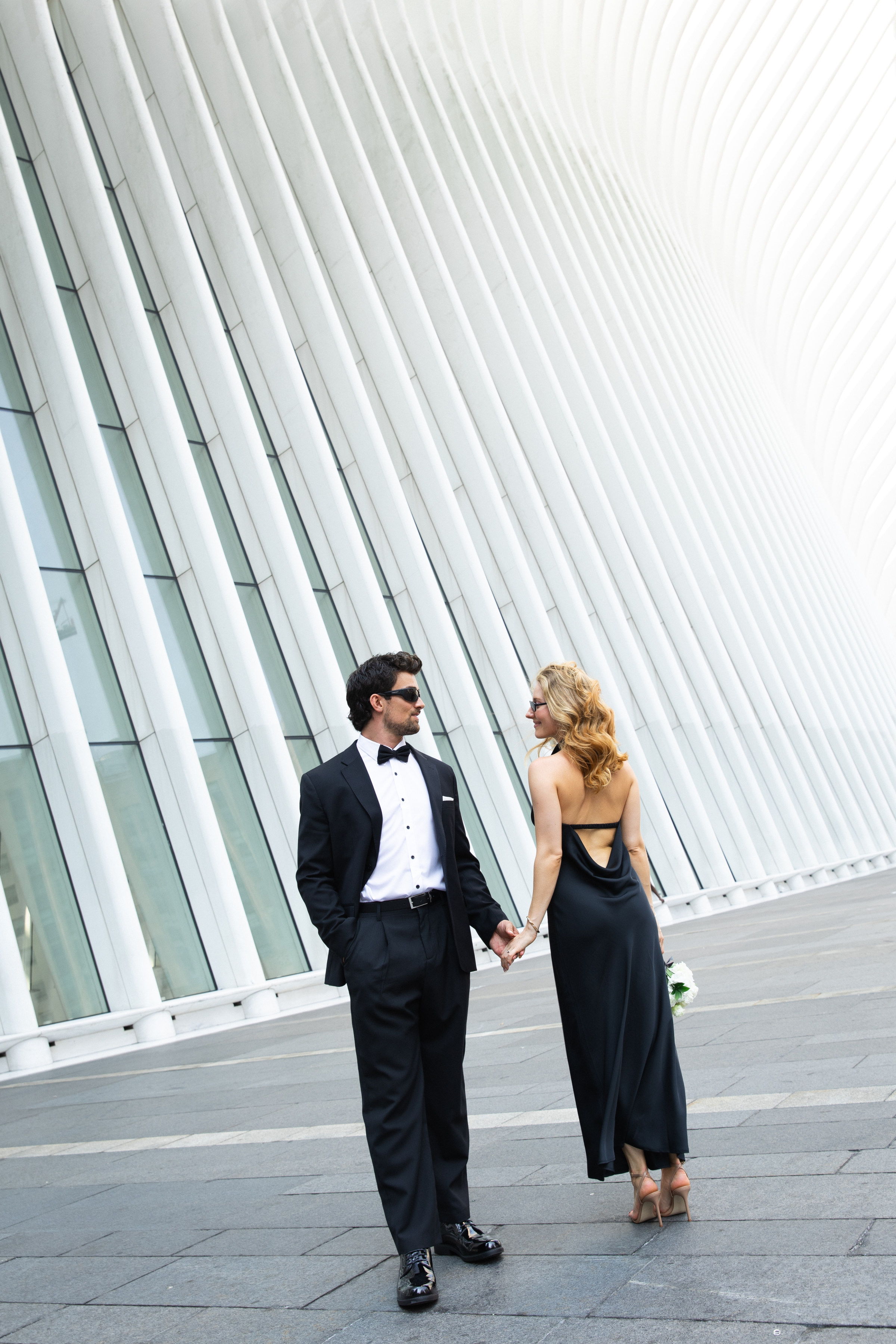 A couple in formal wear holding hands in front of the white ribbed columns of the Oculus in Lower Manhattan