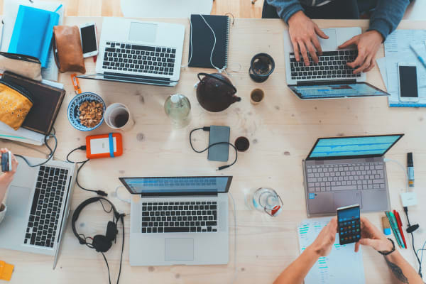Photo of a wooden desk with many people sitting around it, working with laptops. Other things are on the desk such as mobile phones, a bowl of snacks, cup of coffee, some headphones, phone charger and a tea pot