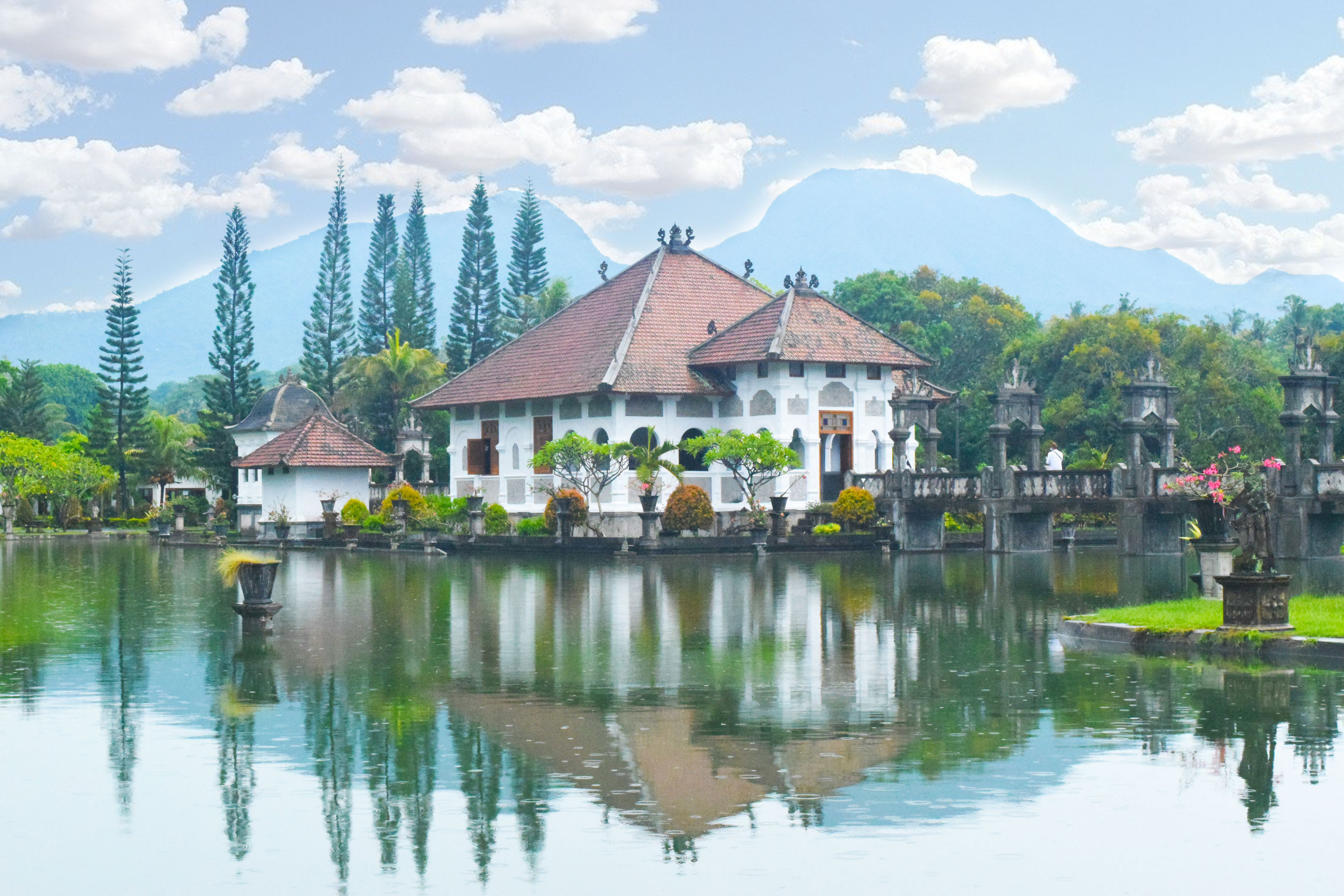 The image showcases the Ujung Water Palace, also known as Taman Ujung, located in Bali, Indonesia. The palace is set against a scenic backdrop of mountains, and it features a large, ornate building surrounded by water gardens. The reflection of the structure in the calm waters adds a tranquil and symmetrical aesthetic to the view. This historical site is a popular destination due to its beautiful architecture and picturesque landscapes.