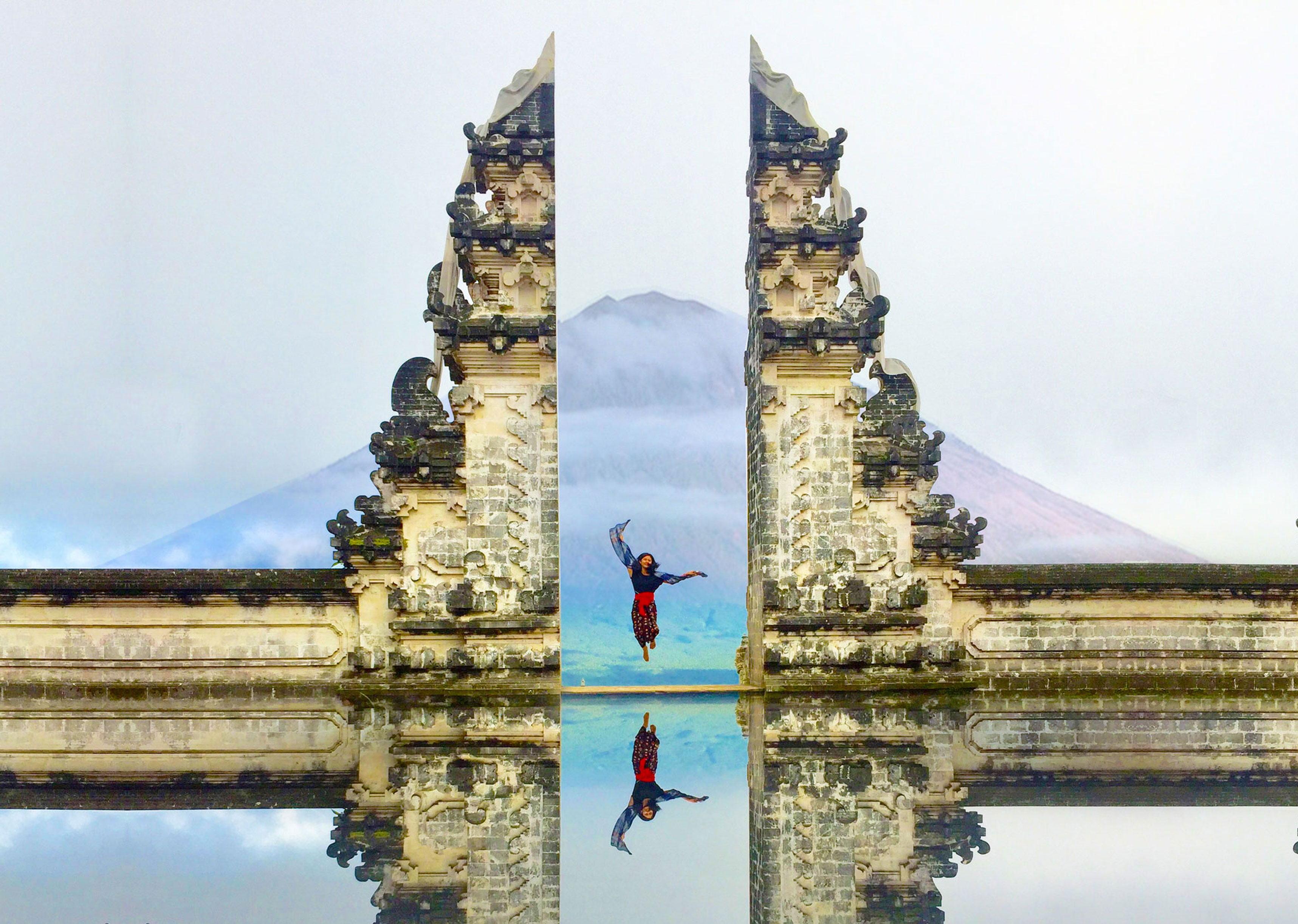 This beautiful image shows the famous Lempuyang Temple gates in Bali, Indonesia. The temple is known for its stunning split gate that perfectly frames Mount Agung, the highest volcano in Bali, which is visible in the background. The person is captured mid-jump, adding a lively and joyful element to the serene landscape. The reflection in the water below the gates creates a symmetric visual effect, making the scene even more picturesque and mesmerizing.