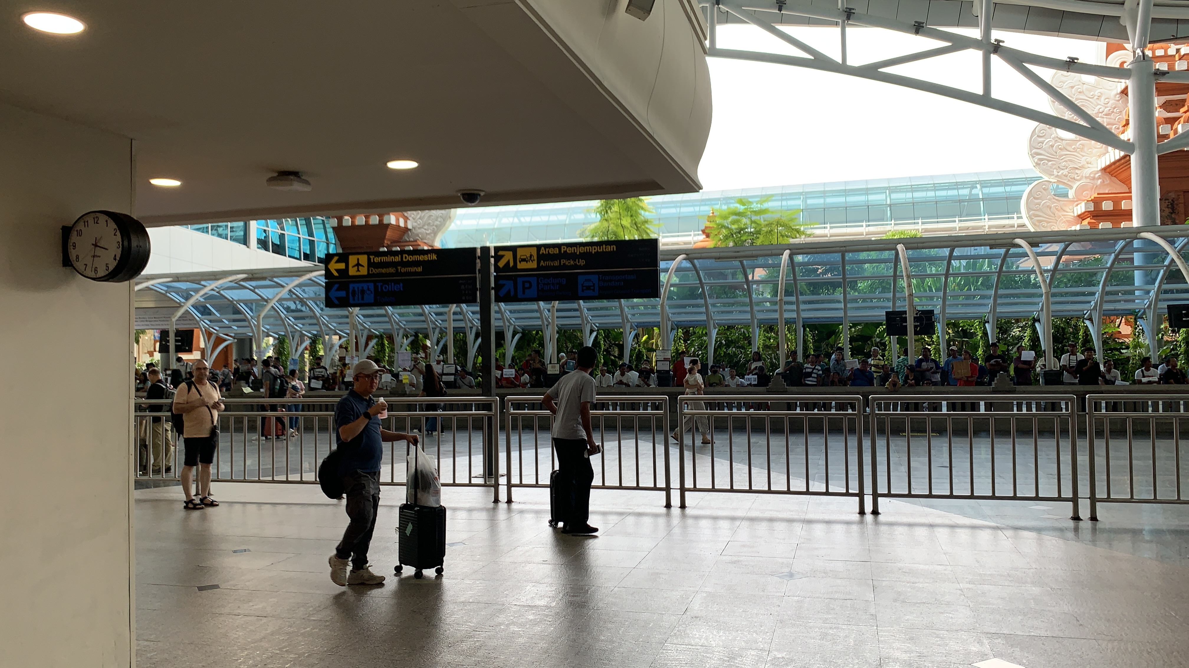 Passengers standing and waiting in the arrival area of Bali's Ngurah Rai International Airport, with directional signs for the Domestic Terminal, Arrival Pick-Up, Building Parking, and Toilets in the background.