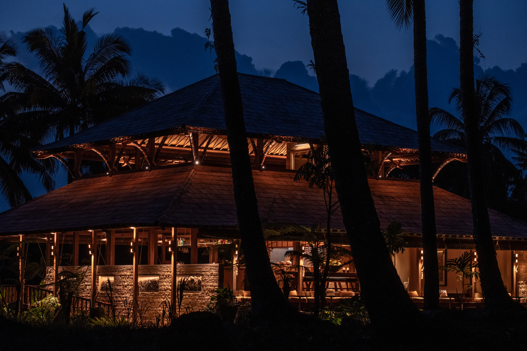 Begawan Biji Restaurant illuminated at night, showcasing its traditional wooden architecture with soft lighting, surrounded by palm trees against a backdrop of twilight sky.