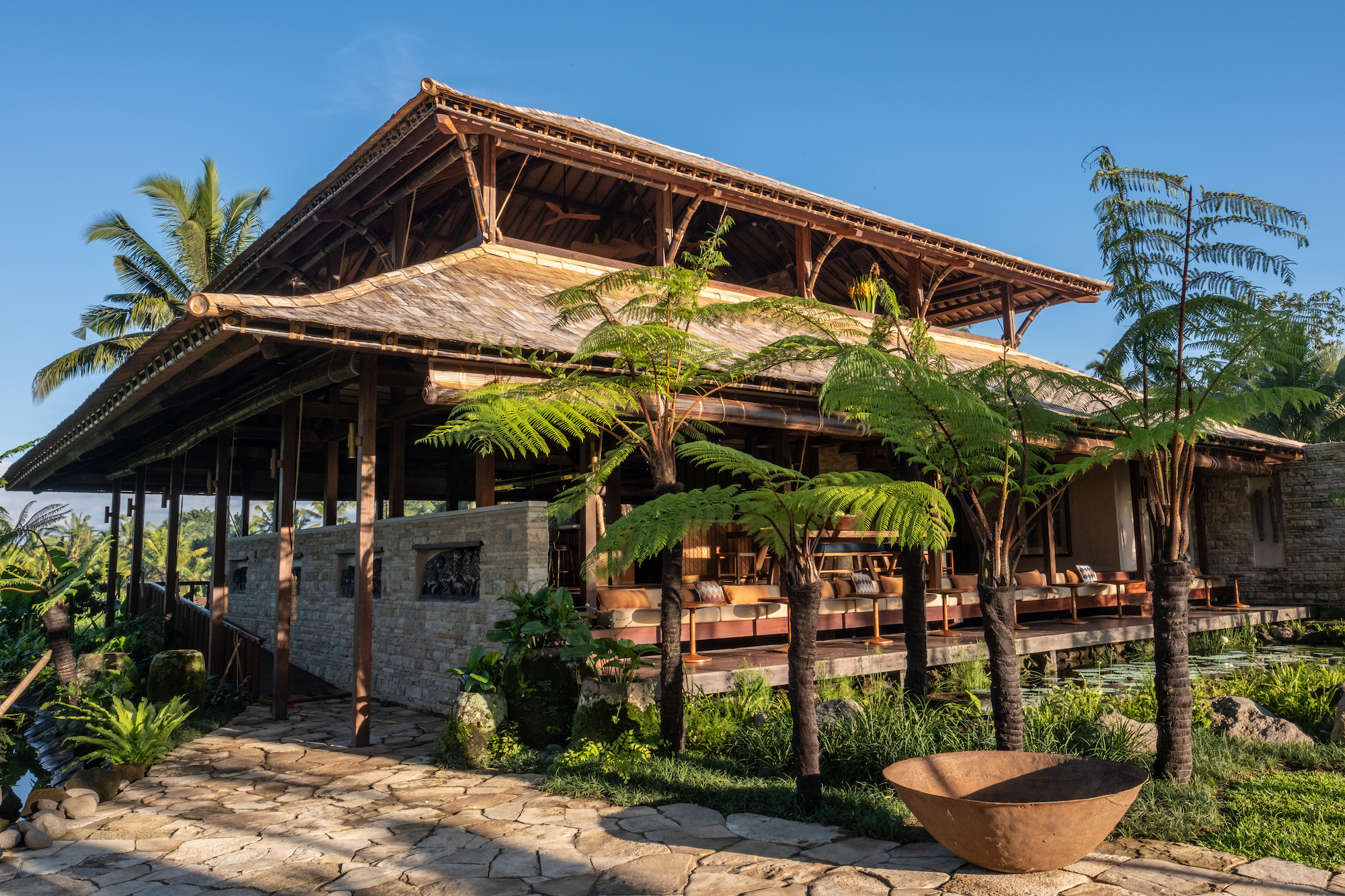 Exterior view of Begawan Biji Restaurant in Bali, featuring traditional architecture with wooden beams, a thatched roof, and lush greenery surrounding the building.