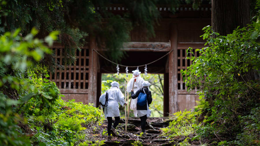 Trois pèlerins en tenue blanche, bâton de bois à la main, passent la porte de bois du mont Haguro
