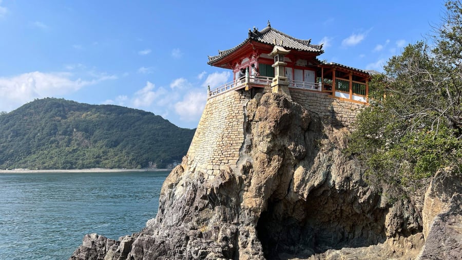 Une salle de temple vermillonne bâtie au sommet d’un rocher qui plonge dans la mer, une île boisée derrière