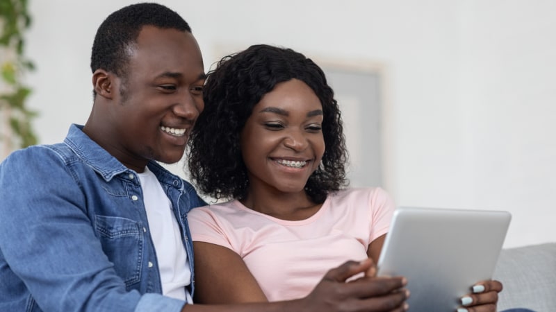 Ein schwarzes Ehepaar sitzt auf einem Sofa und nutzt gemeinsam ein digitales Tablet
