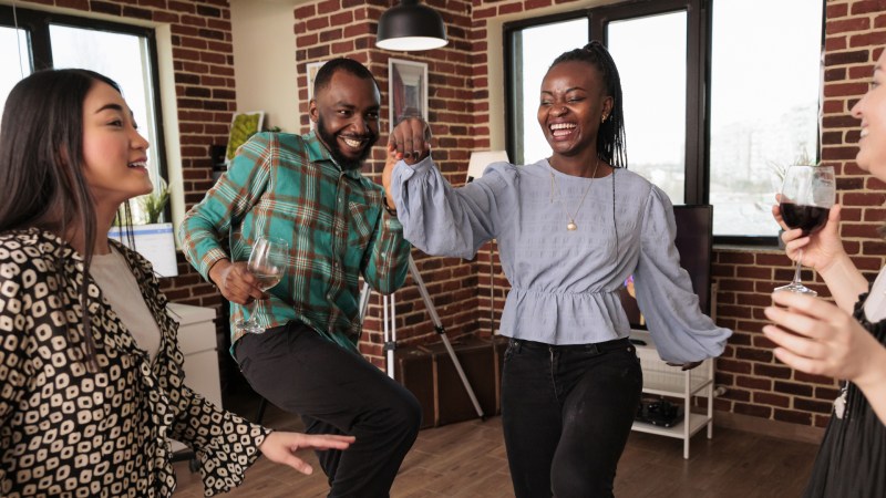 Eine junge afroamerikanische Frau tanzt und lächelt inmitten einer Gruppe von Menschen unterschiedlicher Herkunft, die lachen und bei einem Treffen in einem Wohnzimmer Wein trinken