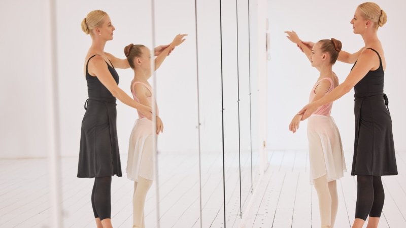 A ballet teacher training a young girl in a dance studio in front of a mirror