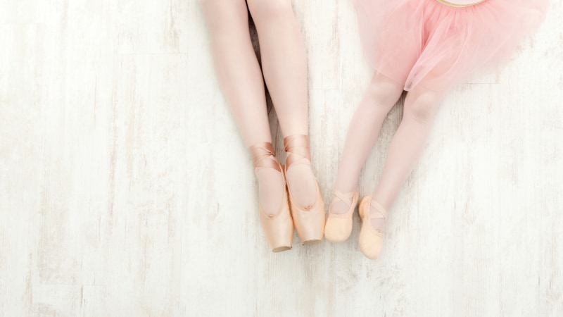 Set fra oven af benene på en lille pige og en ung ballerina i tåspidssko på et hvidt trægulv med fri plads