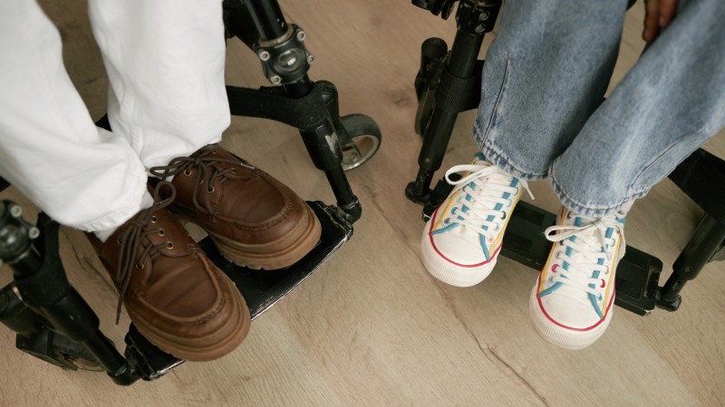 Füße eines Mannes in formellen Schuhen und einer Frau in Turnschuhen, die in Innenräumen in Rollstühlen sitzen