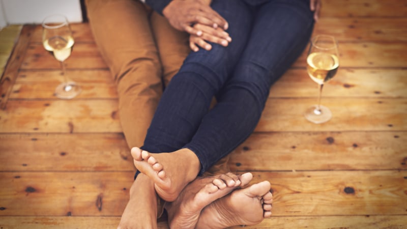 Ein Paar sitzt mit Champagnergläsern auf einem Holzboden und zeigt die Beine der beiden Personen in einer Wohnung