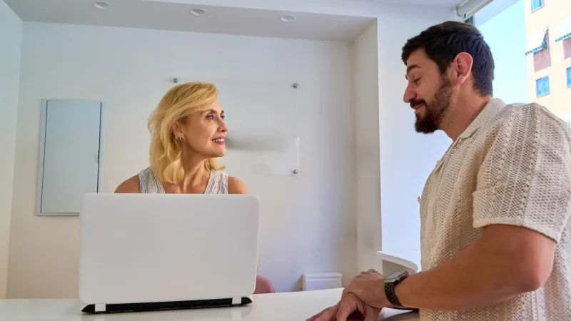 Eine Frau und ein Mann unterhalten sich an einem Empfangstresen mit einem Laptop.