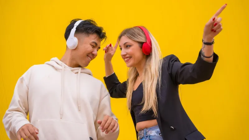 Ein asiatischer Mann und eine weiße Frau tanzen mit Kopfhörern Bachata und Salsa vor gelbem Hintergrund