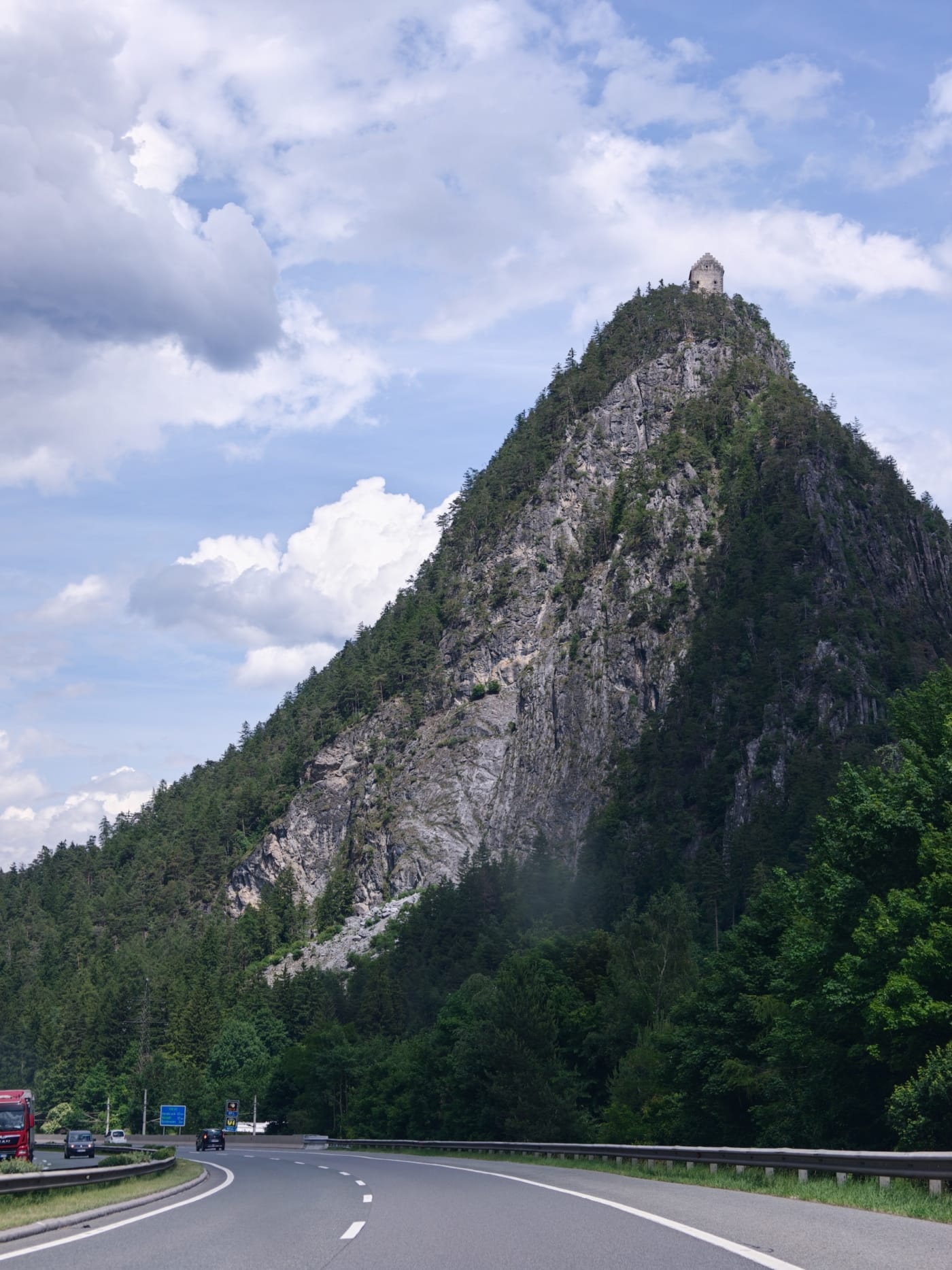 Rocky forested peak with a small stone tower at the summit above a curving mountain road