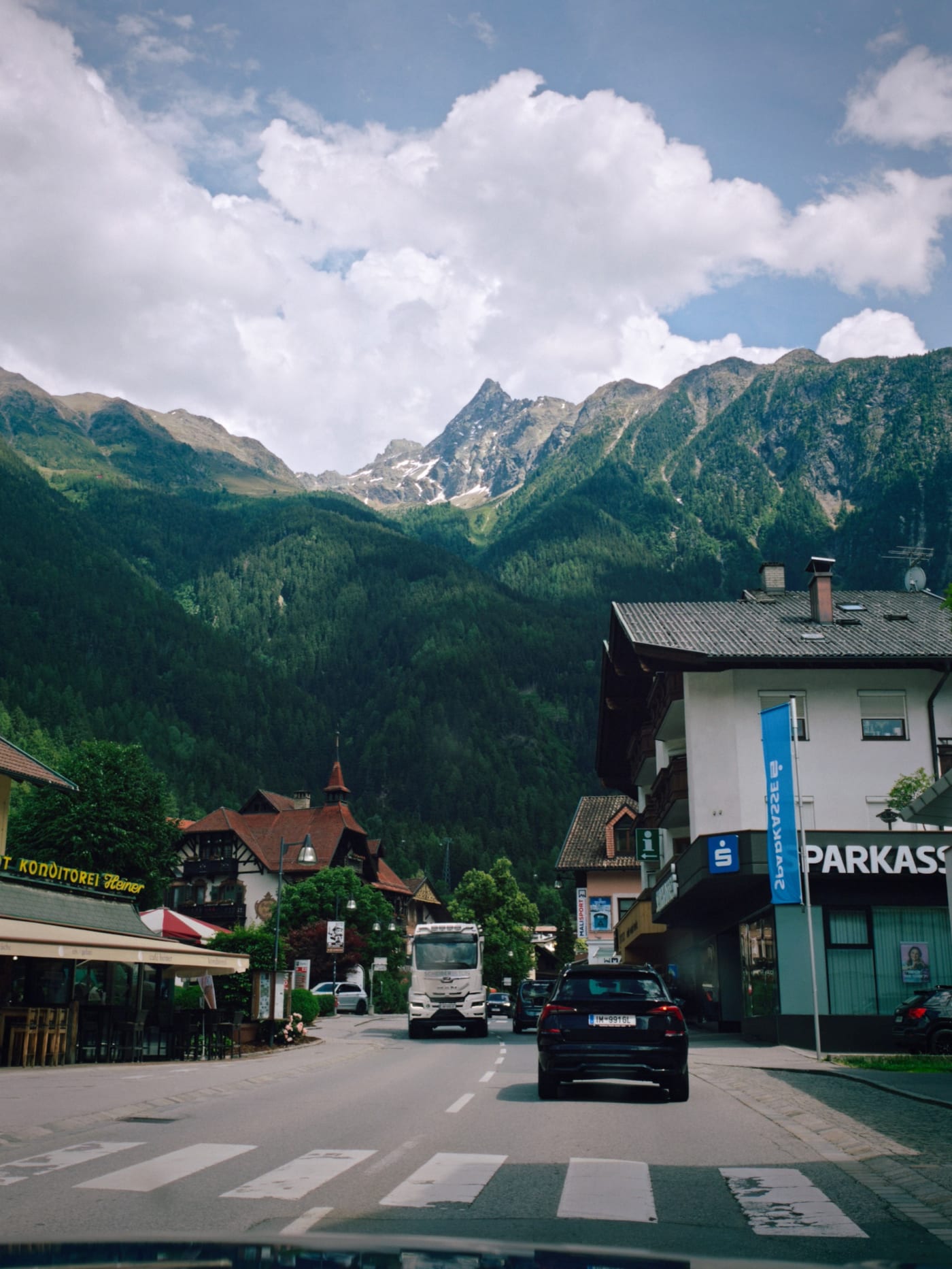 Alpine village street with cars and shops below high green mountains and bright clouds