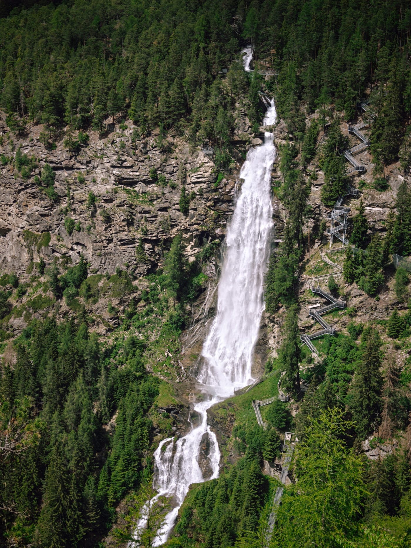 Tall waterfall dropping through a forested rocky cliff with stairways along the slope