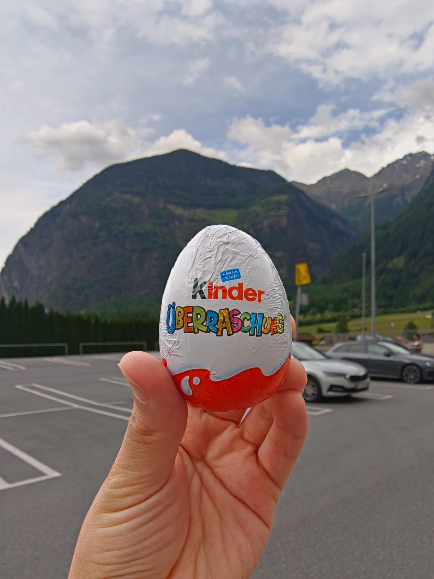 Hand holding a Kinder Surprise egg in a mountain parking lot with alpine peaks in the background