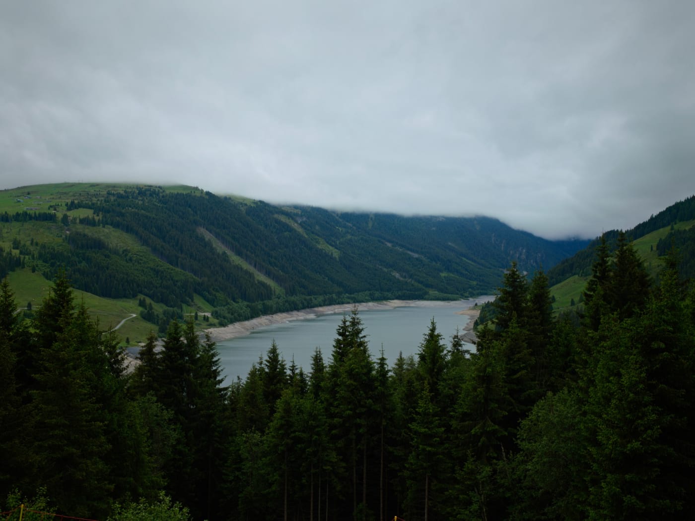 Wide view of a pale alpine lake surrounded by dark evergreen forest and cloud-covered hills