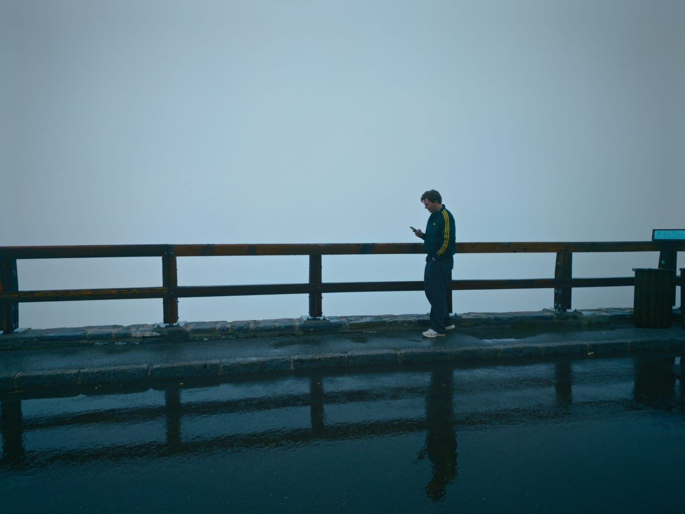 Person standing by a wooden guardrail on a foggy overlook with reflections on wet pavement