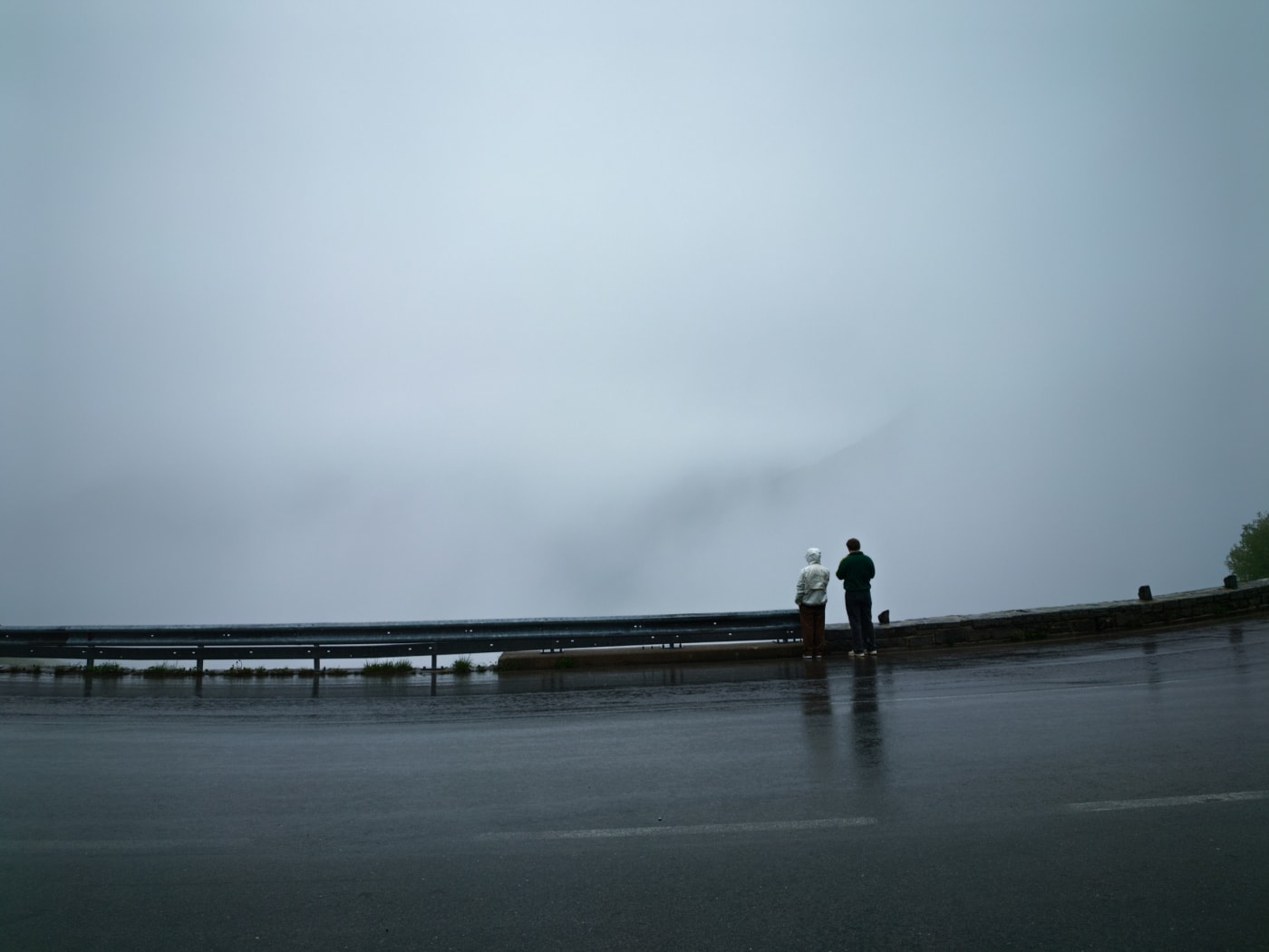 Two people standing at a rainy roadside viewpoint looking into dense fog