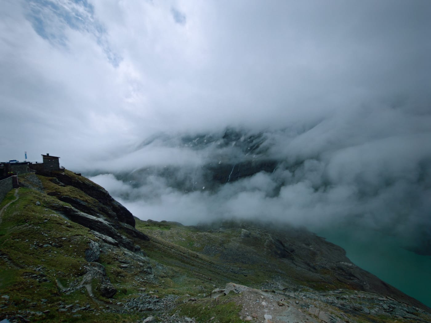 Clouds wrapping around a mountain ridge above a turquoise alpine reservoir