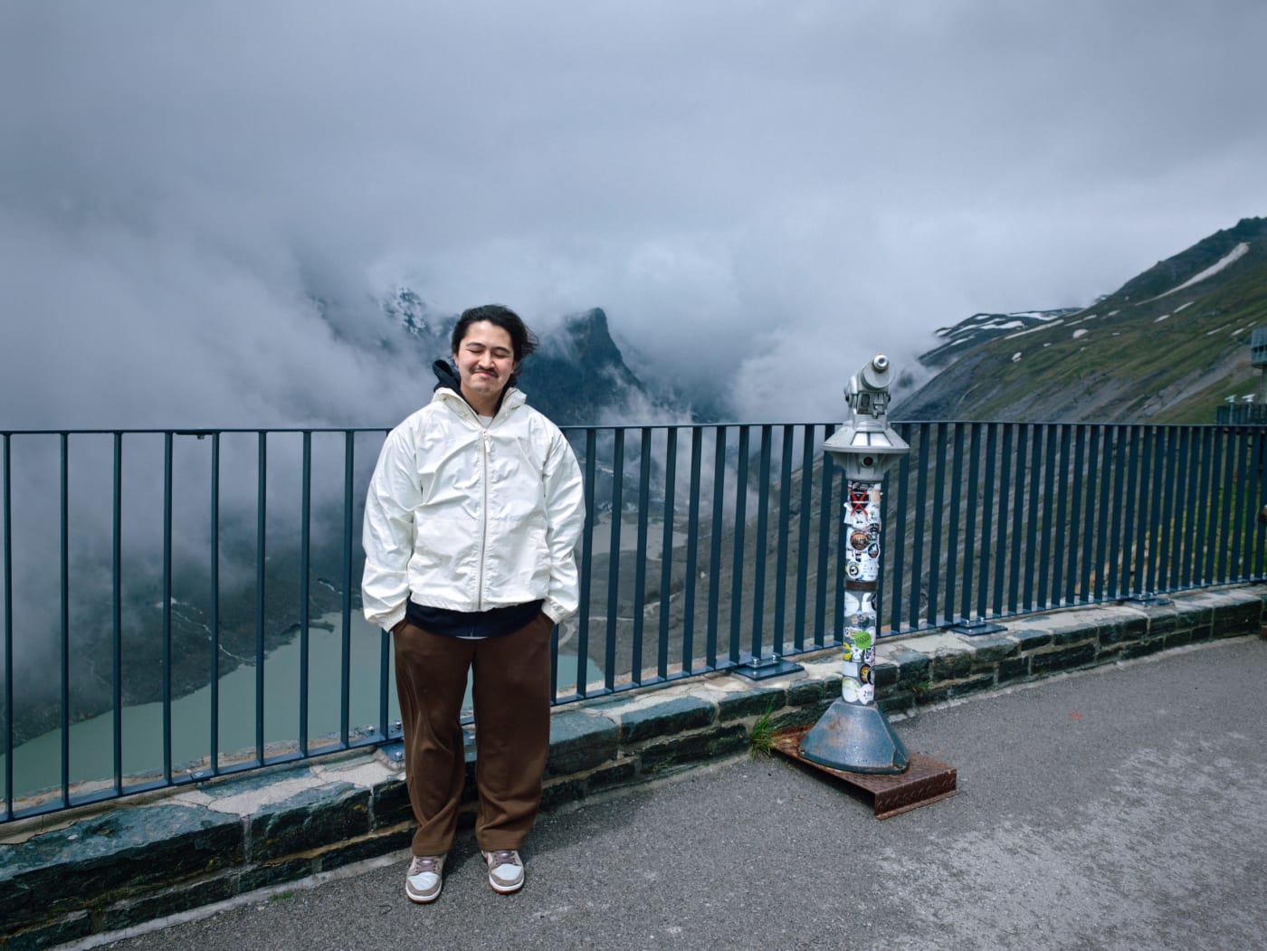 Person in a white jacket standing beside a coin-operated telescope at a misty mountain overlook
