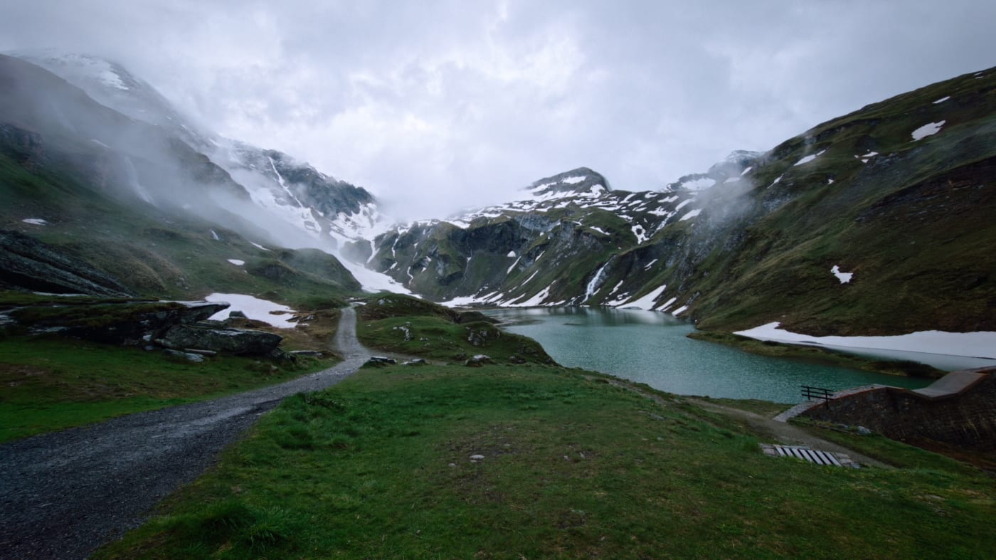 Wide panorama of a turquoise alpine lake surrounded by snowy green mountains and low clouds