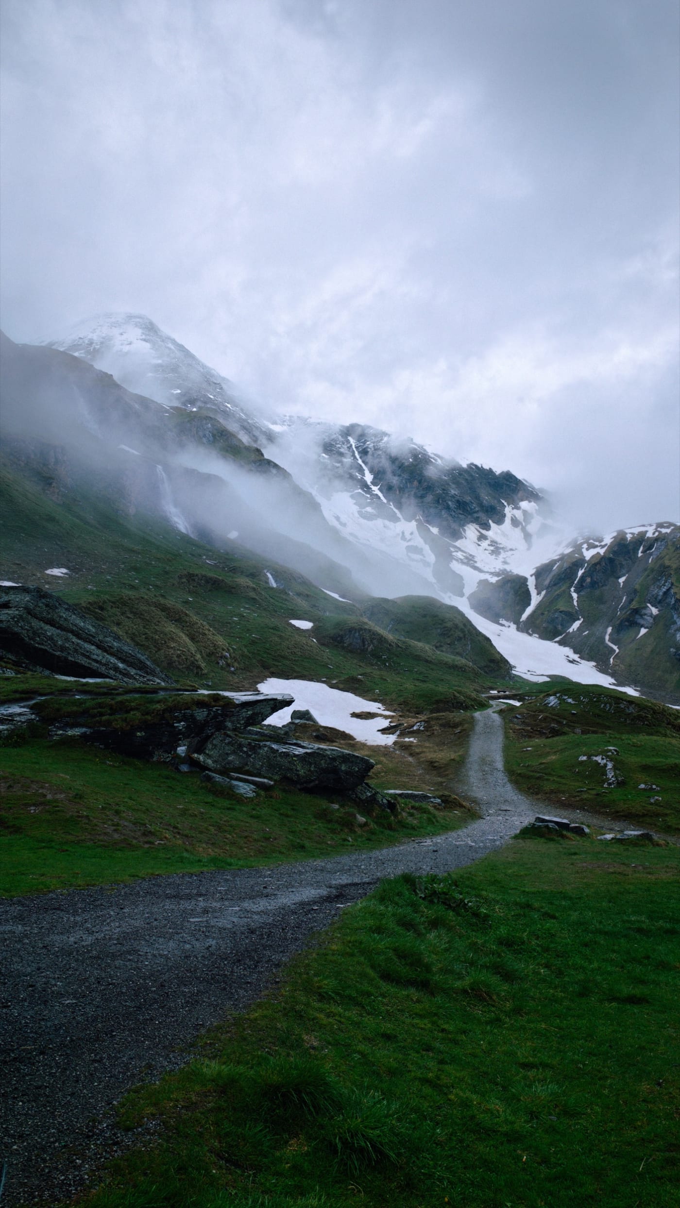 Gravel path leading toward snowy alpine slopes and clouds in wet weather