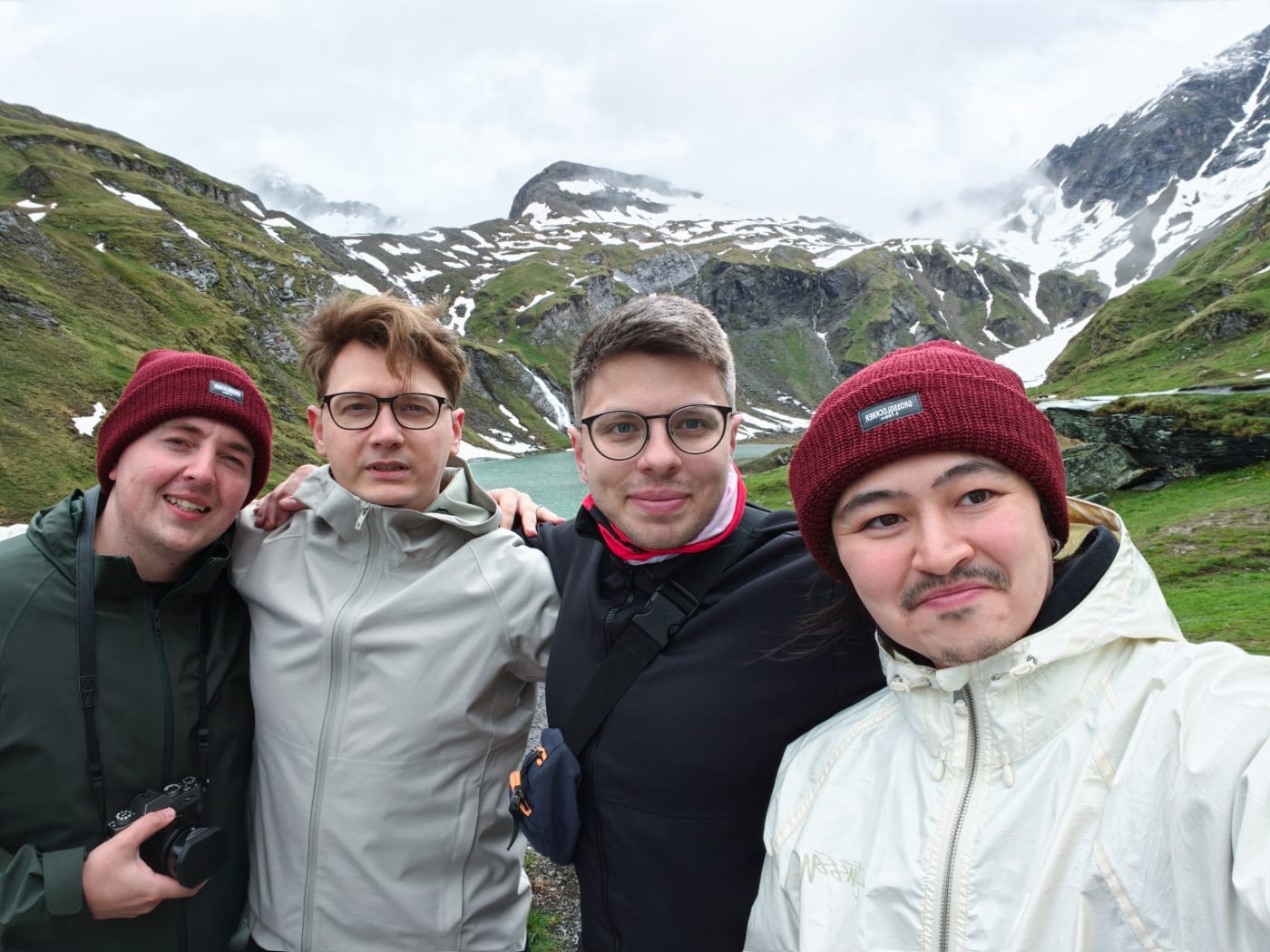 Four friends taking a selfie in front of an alpine lake and snow-patched mountains