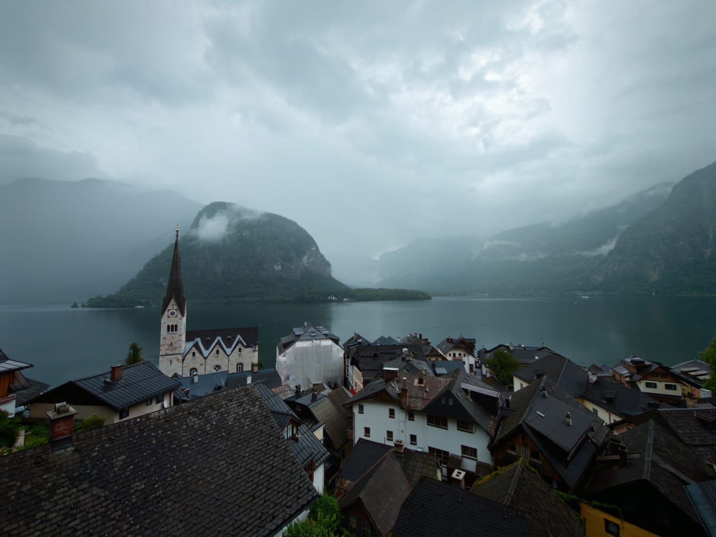 Rainy view over Hallstatt rooftops, church, lake, and mist-covered mountains