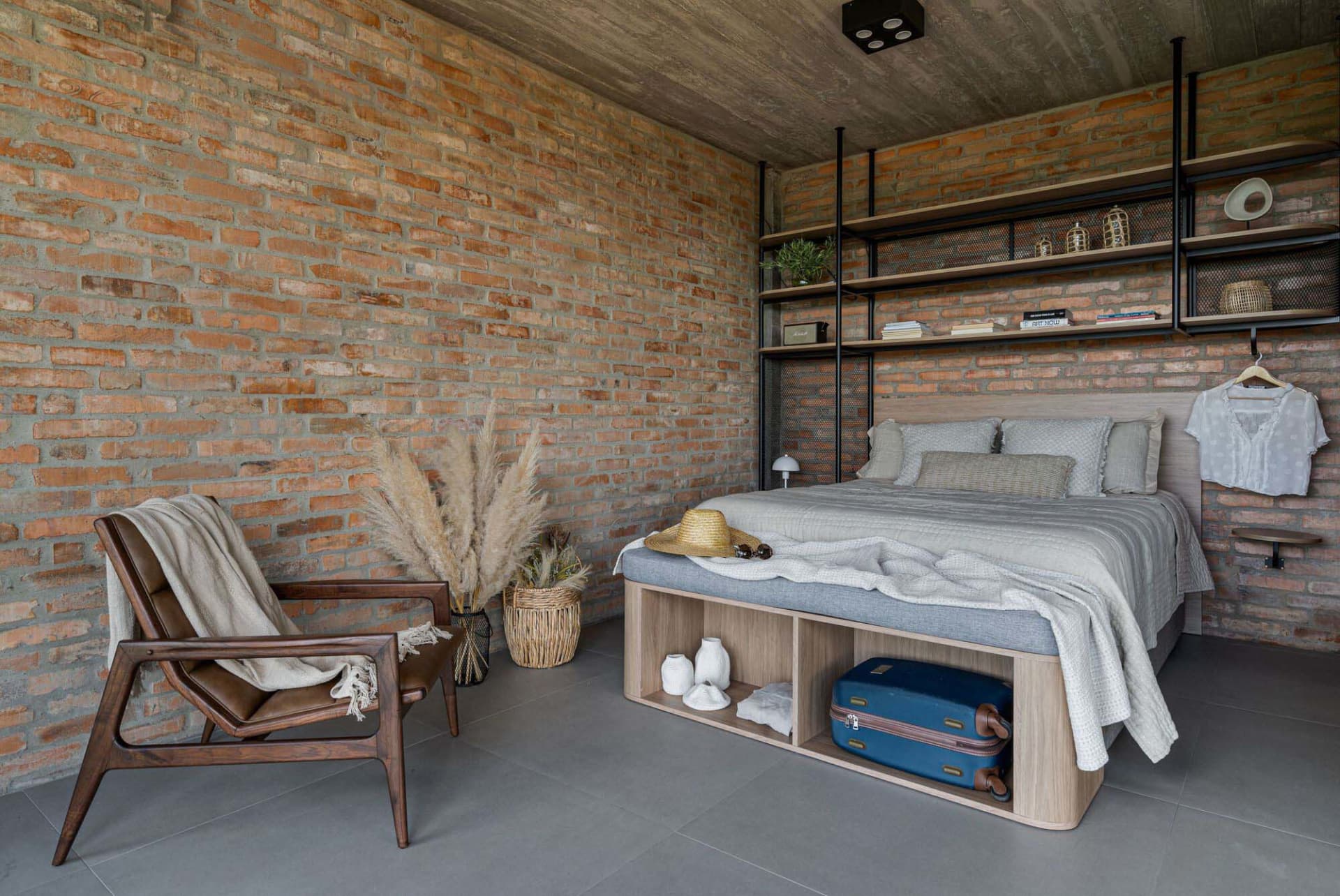 A modern, minimalist bedroom featuring brick and concrete walls, a wooden bed frame, and a leather chair.