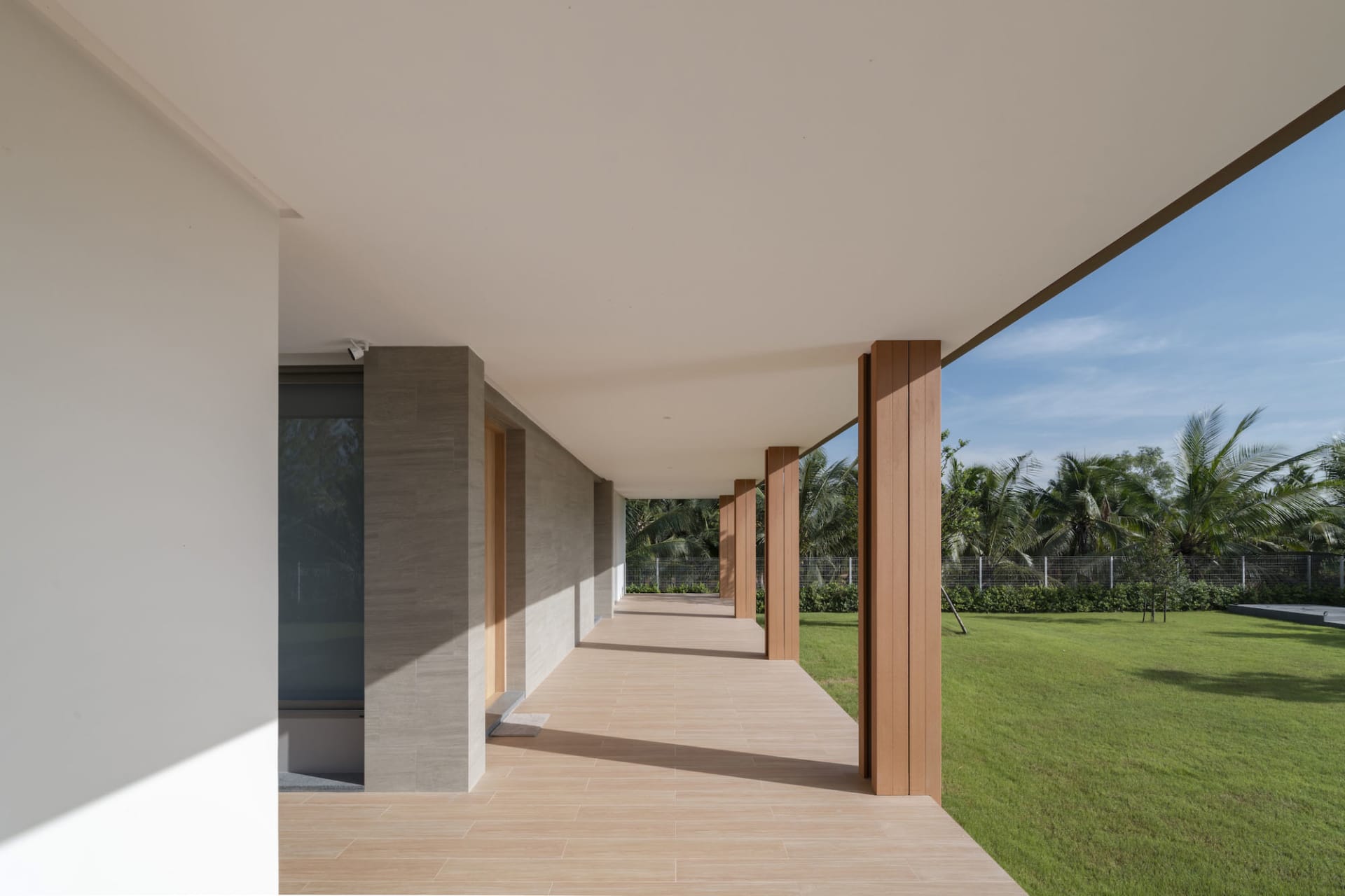 A modern architectural walkway with wooden pillars and concrete walls, surrounded by greenery and blue sky.