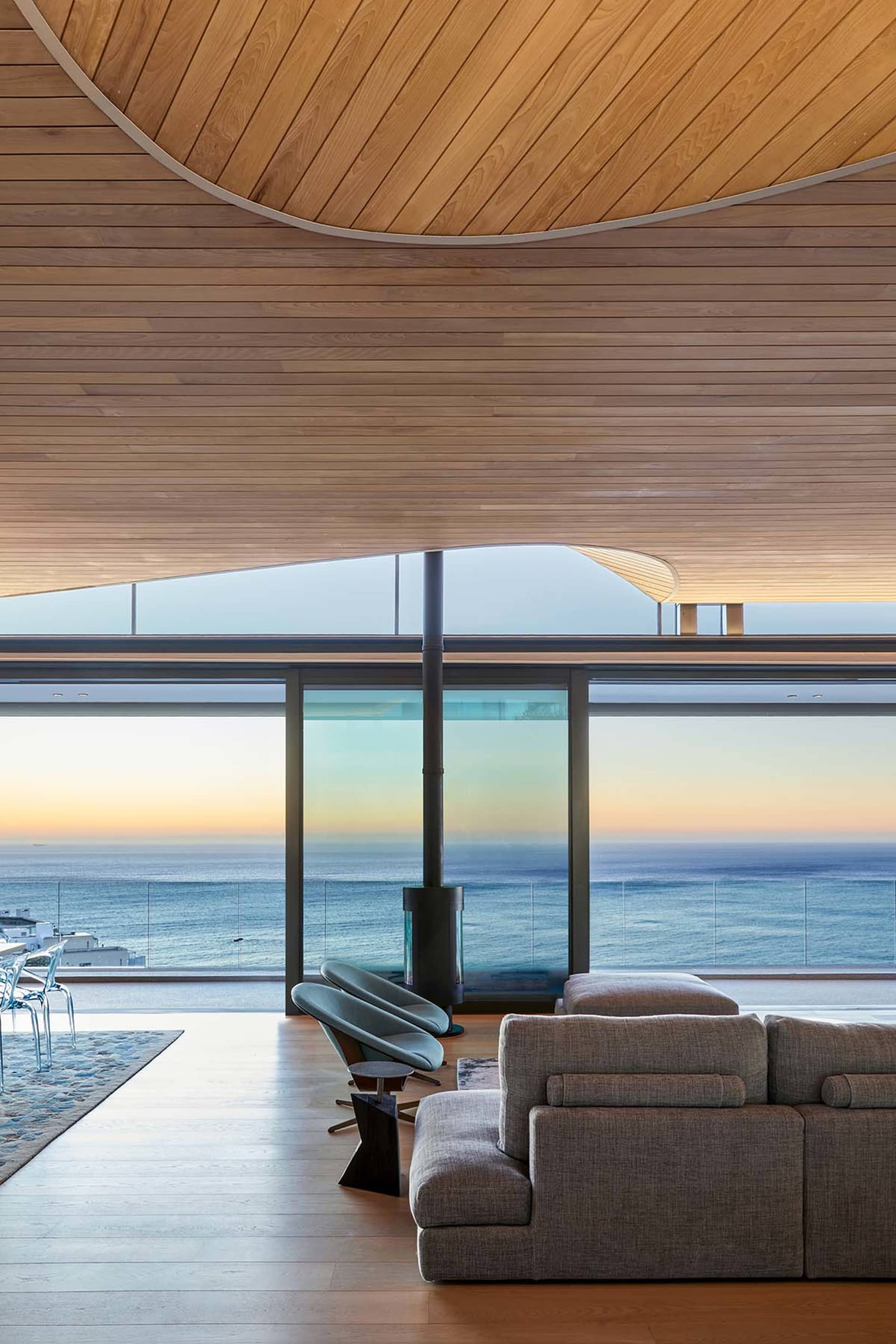 Modern minimalist living room with wood furniture and floor-to-ceiling windows overlooking the ocean.