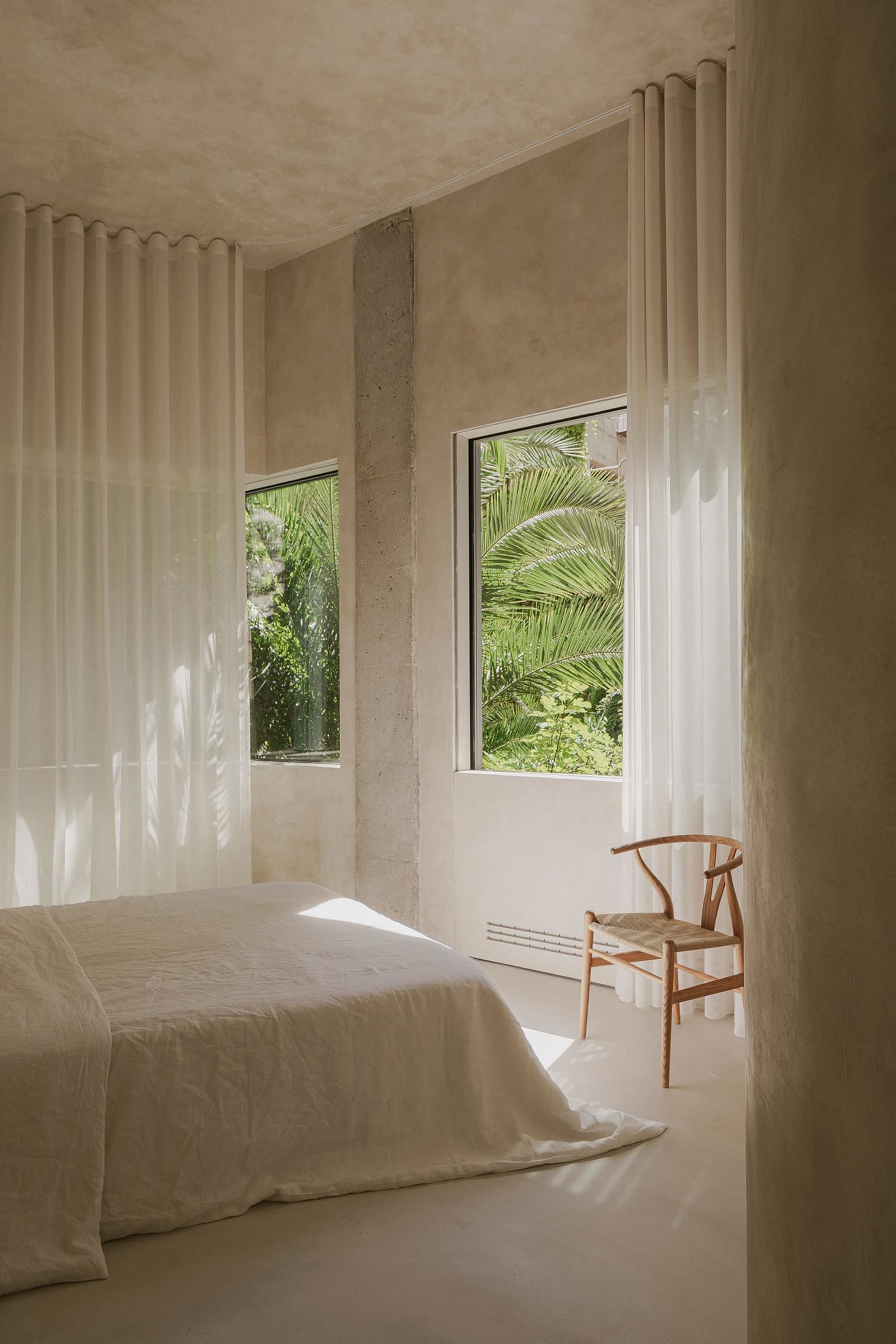 A minimalist bedroom with natural fibers, featuring a bed with white sheets, a wooden chair, and sheer curtains.