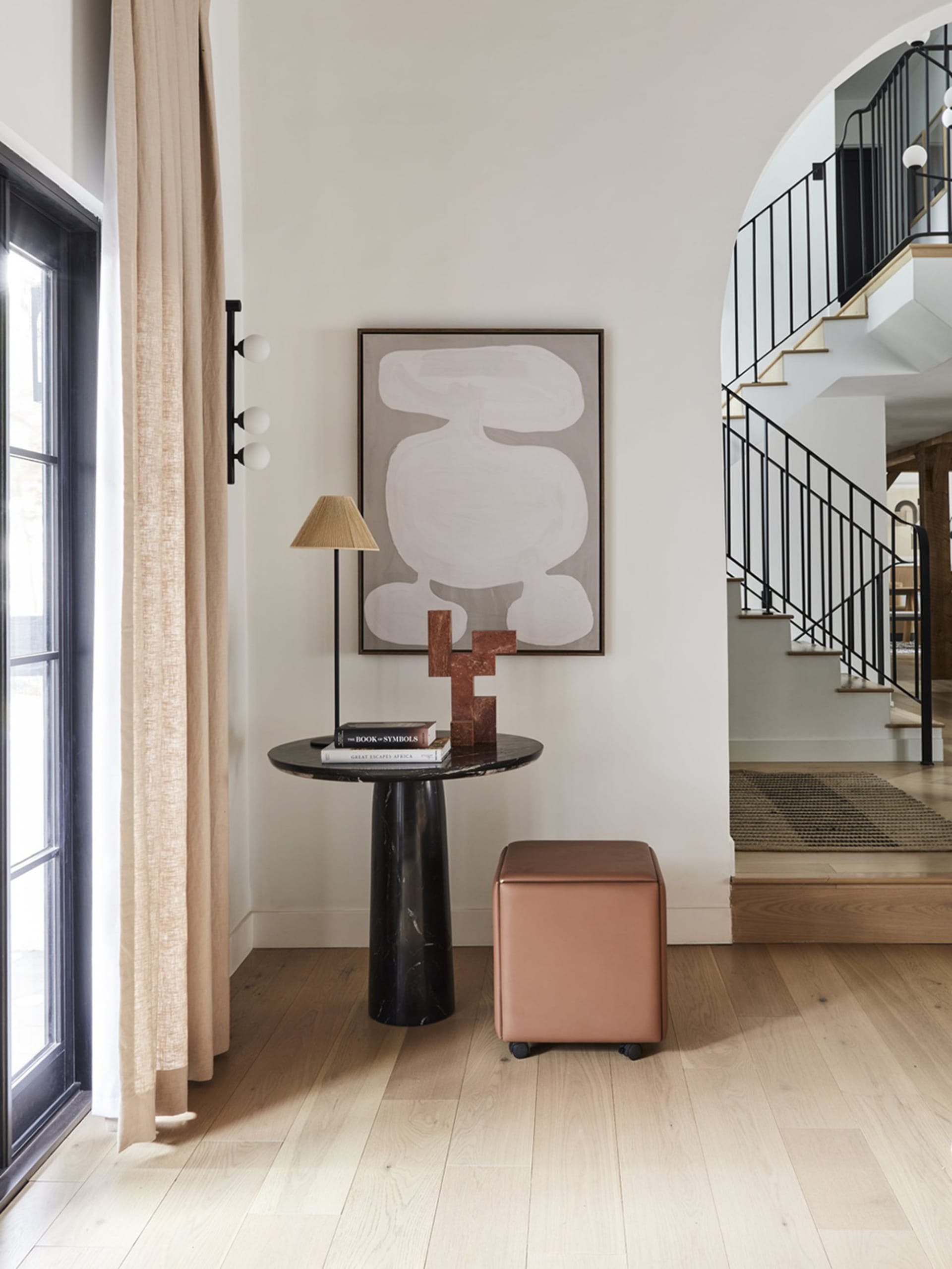 A minimalist entryway with a black marble pedestal table, a pink leather cube ottoman on wheels, and a modern light fixture w