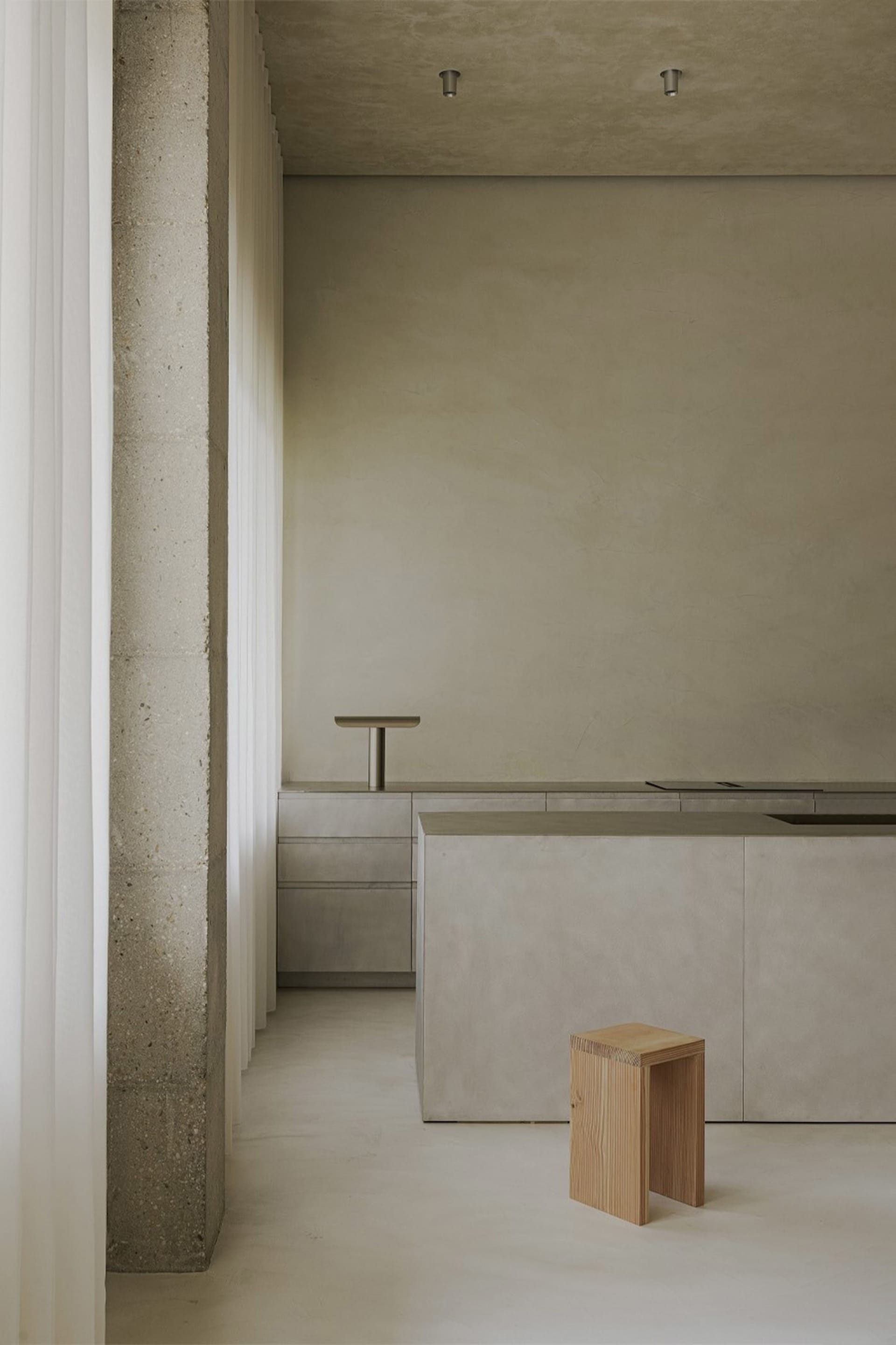 A modern, minimalist kitchen with concrete walls, countertops, and a wooden stool.