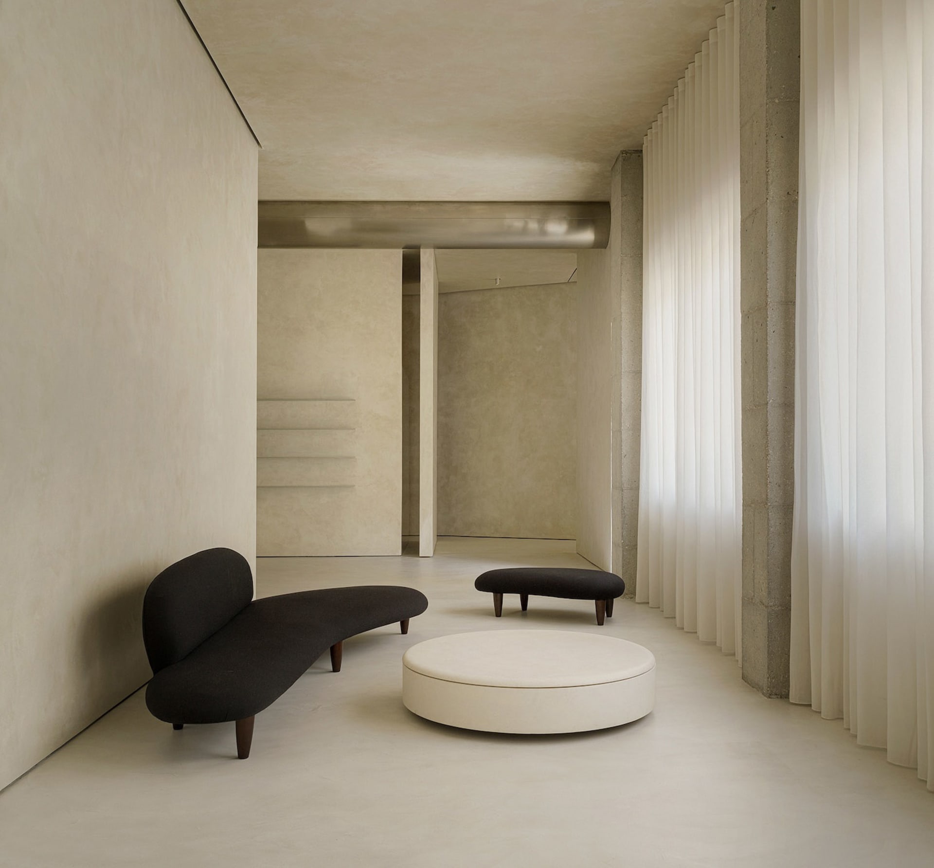 A minimalist living room featuring a black curved sofa, a round white ottoman, and a concrete floor.