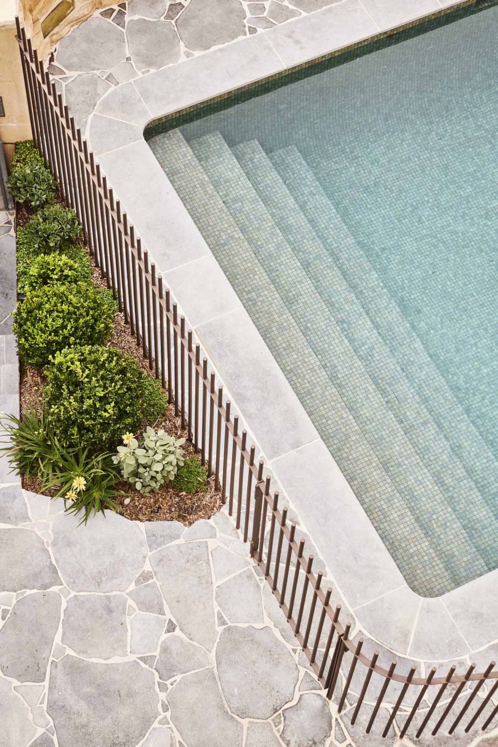 A modern minimalist pool with blue water, surrounded by stone flooring and a metal railing, and adorned with green plants.