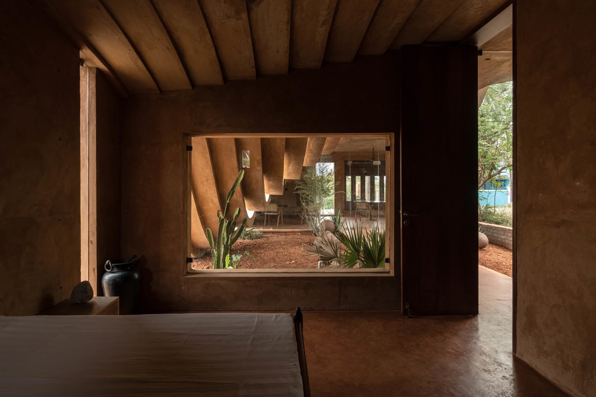 A modern bedroom featuring textured clay walls, concrete floors, and a window overlooking a courtyard with plants.