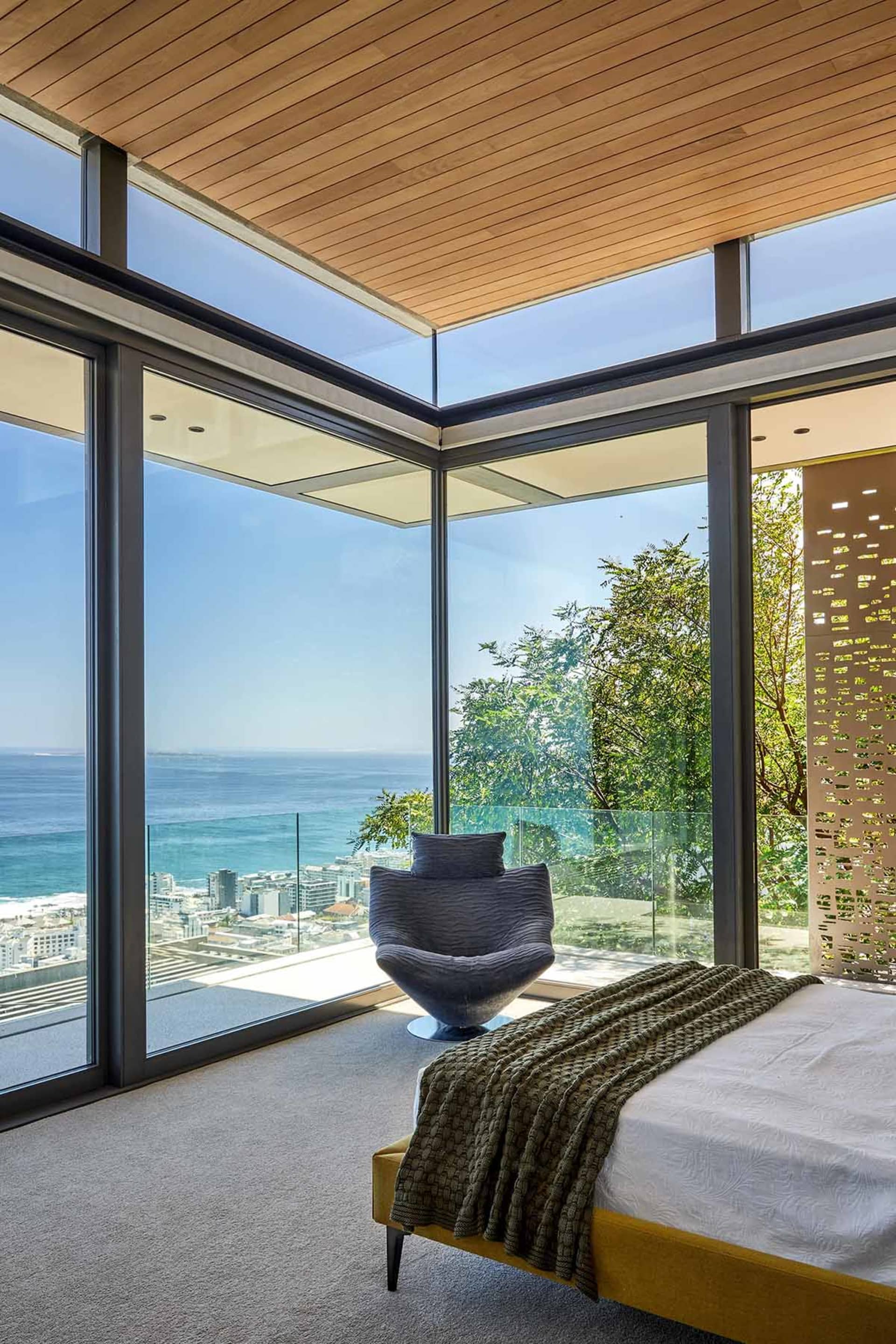 Modern residential bedroom with a glass wall overlooking the city and the ocean, featuring a cozy seating area and a comforta