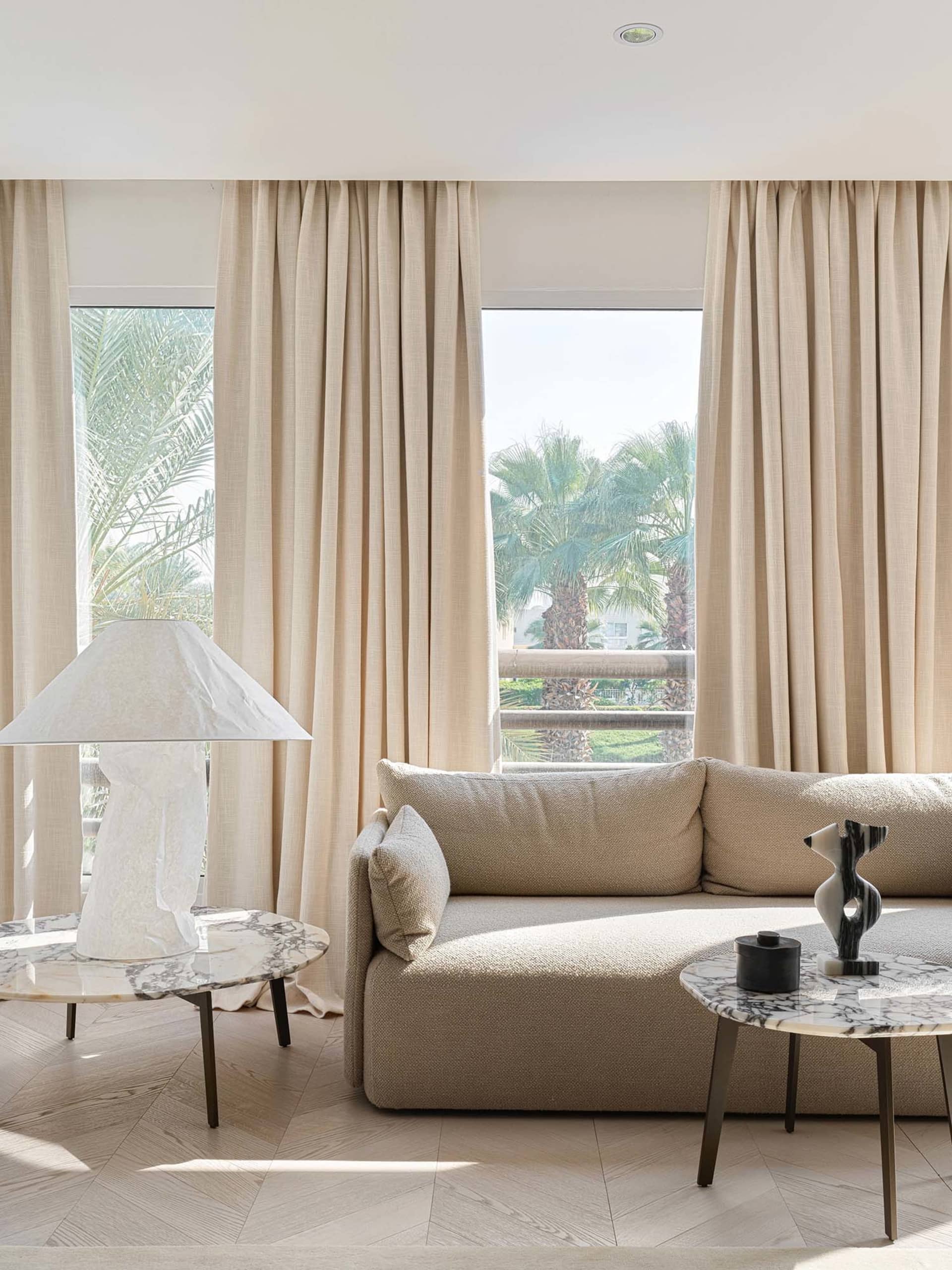 Modern living room featuring a fabric sofa and marble tables with a view of palm trees outside.