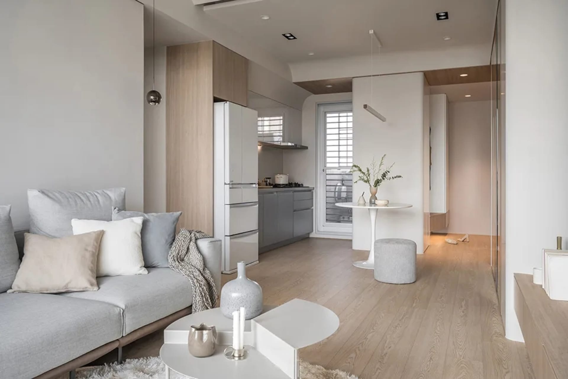 A minimalist living room with a grey sofa, wooden flooring, and a kitchen area in the background.
