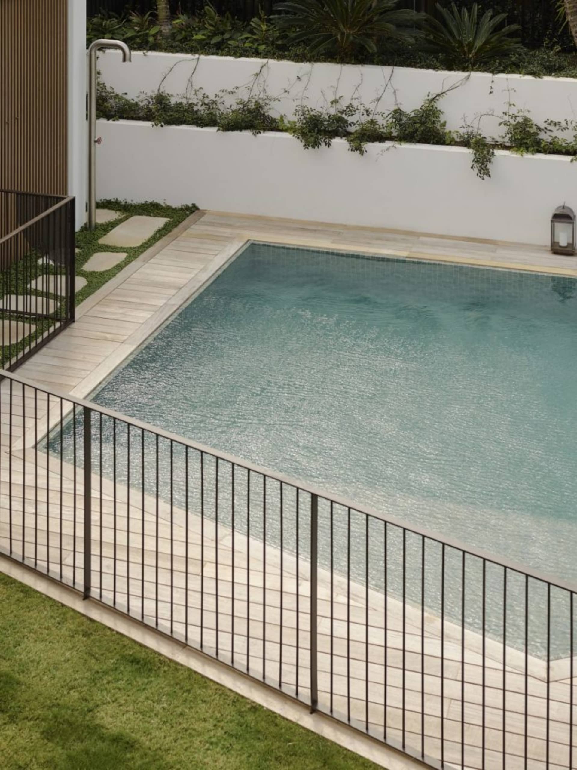 A modern minimalist swimming pool with glass walls and a wooden deck surrounded by greenery.