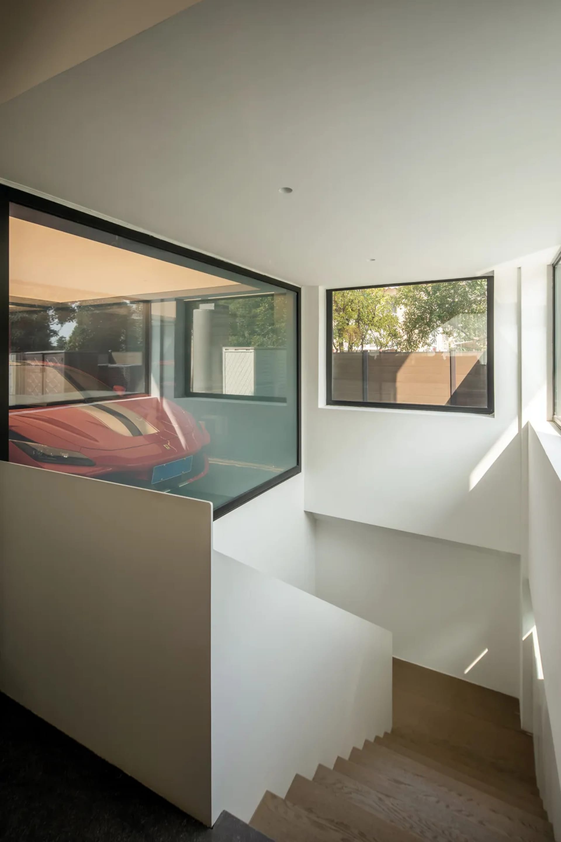 Modern residential staircase with white concrete walls and floor, featuring a large window revealing a sleek red sports car p