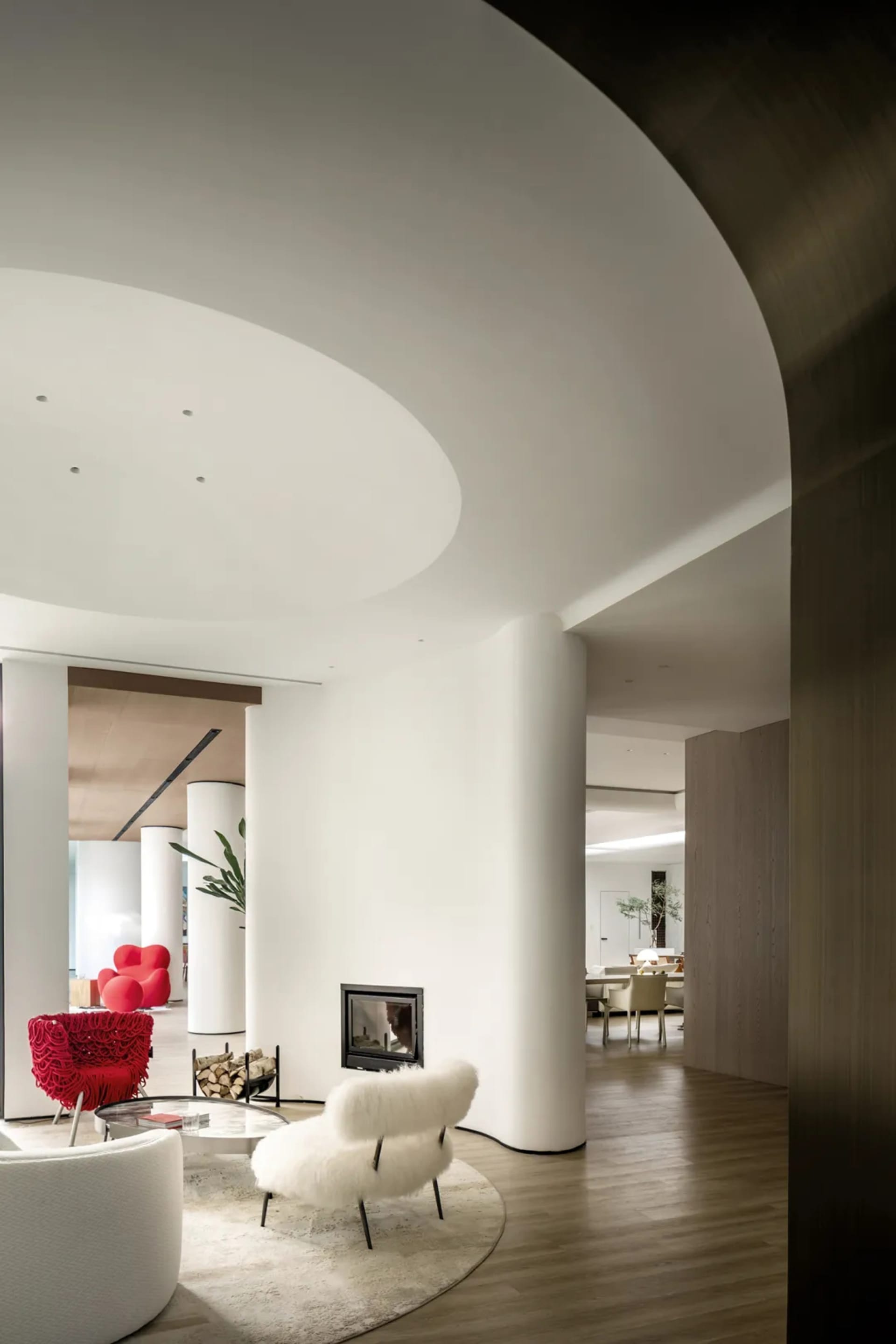A modern living room with a white fur-covered chair, a round glass table, and a fireplace. The space features clean lines, mi