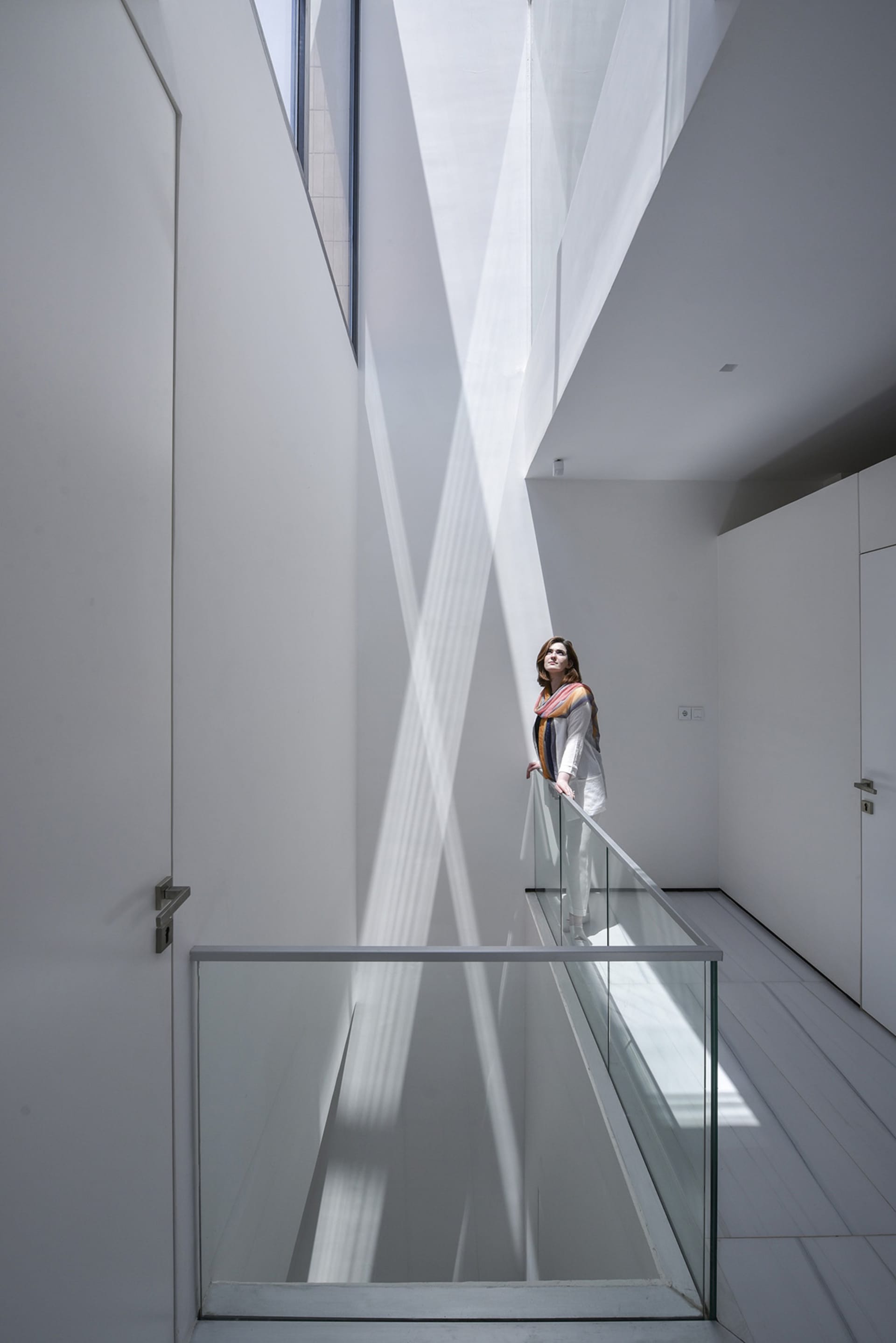 A woman stands on a glass wall railing in a modern, white interior space.